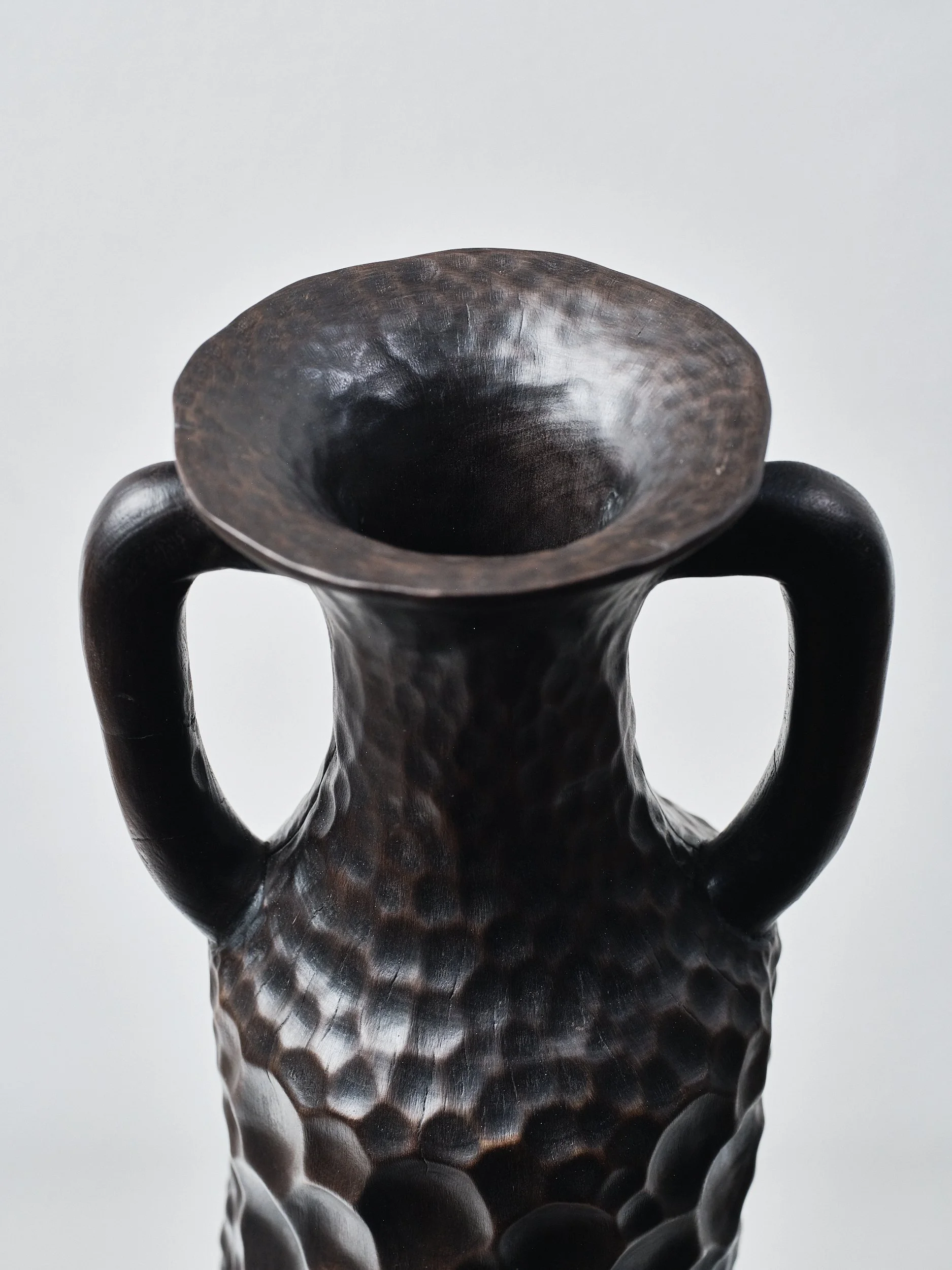Top detail of a black sculpture of Greek Amphora with polished honeycomb textured pattern. Shows a wide opening with two handles is shown against a plain light background.