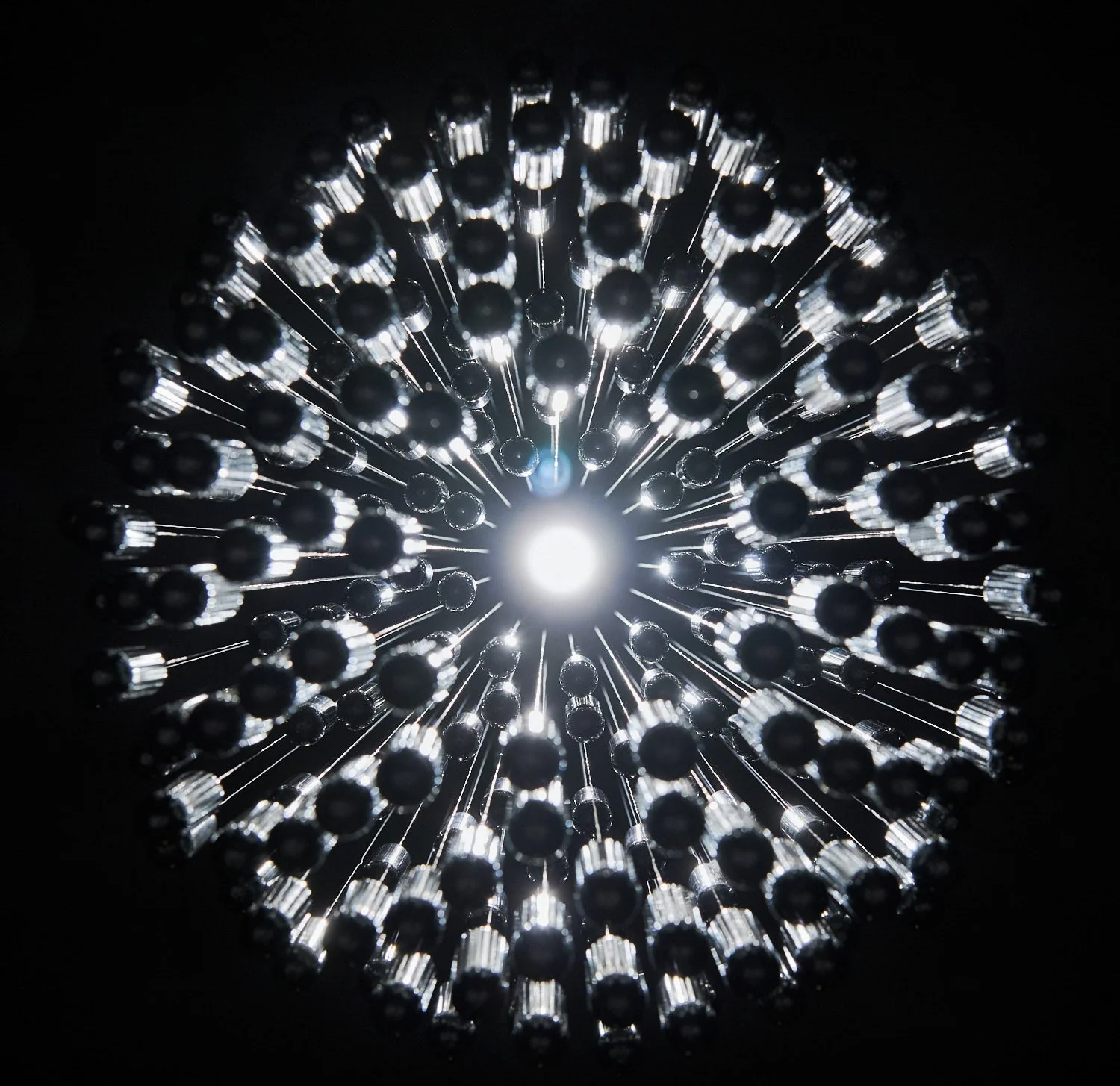 Close-up of the underside of the NOS Sphere chandelier with multiple polished silver nitrous oxide canisters arranged in a circular pattern, glowing against a dark background.