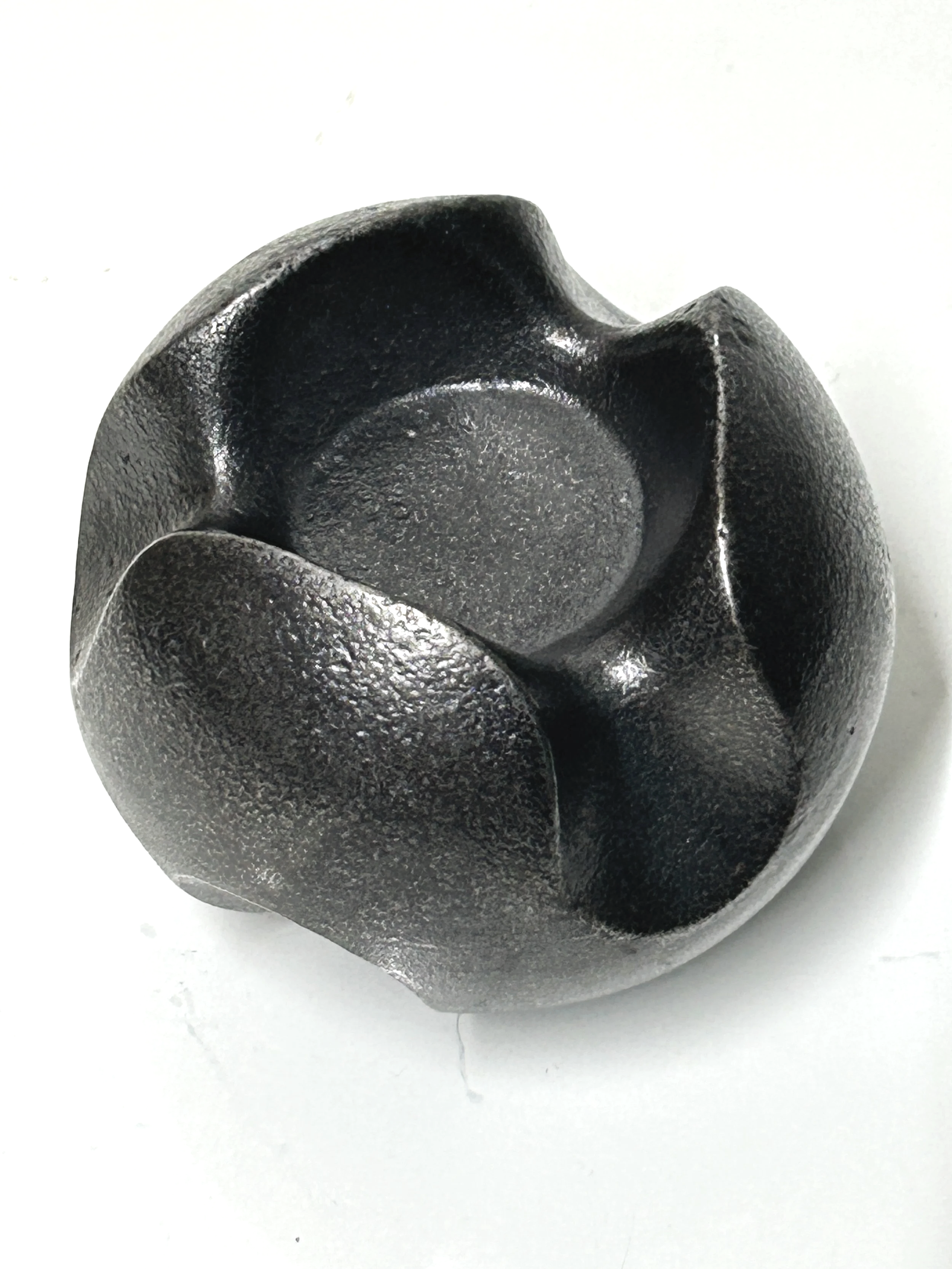 Close-up of a blackened and polished cast iron sculpture, resembling a spherical mechanical part with three evenly spaced notches around its circumference, placed on a white surface.