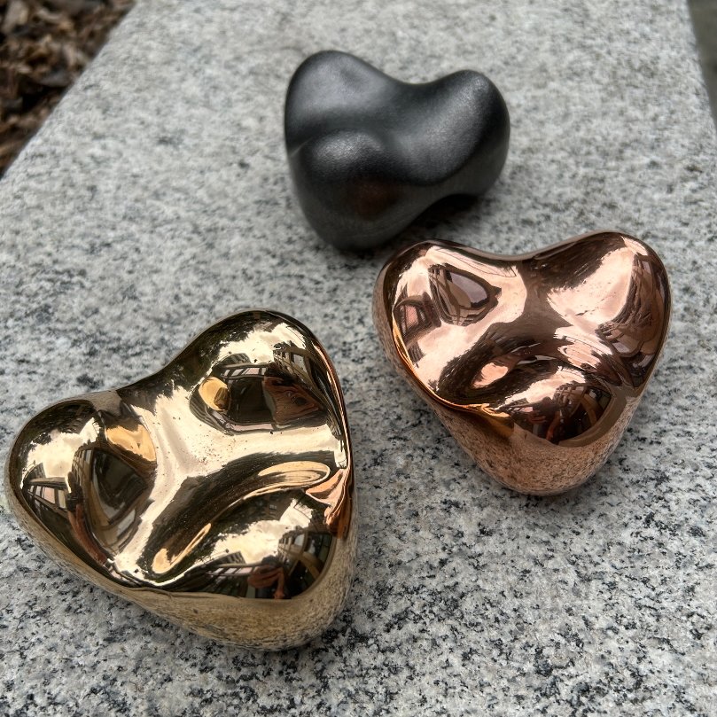 Three polished pebble like art objects called Puck. Two are made in polished bronze and copper, and one is matte black rumbled cast iron, resting on a rough grey stone surface.