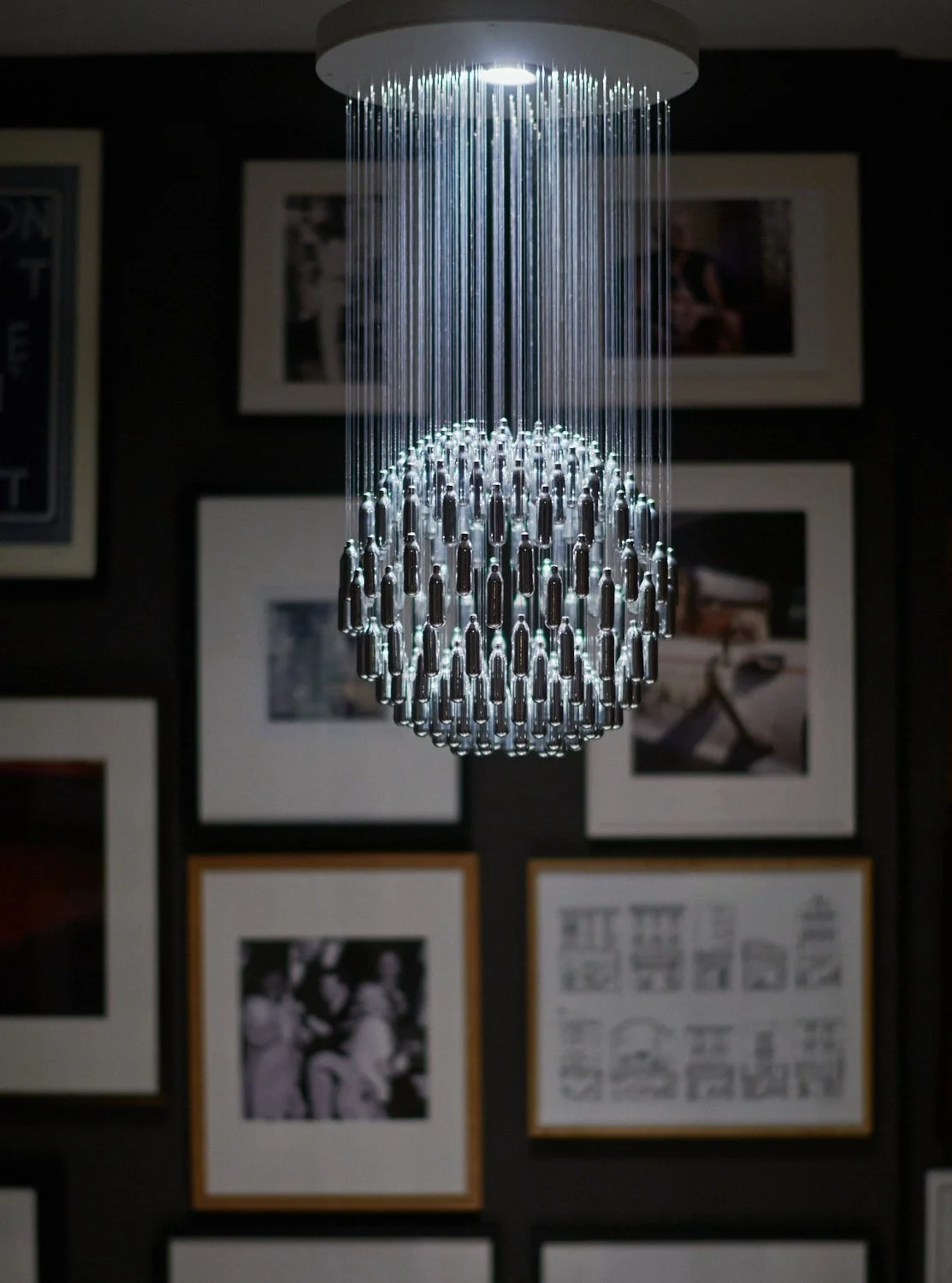 Sustainably designed NOS Sphere 228 chandelier, made from 228 recycled found nitrous oxide canisters in front of a wall of framed photographs