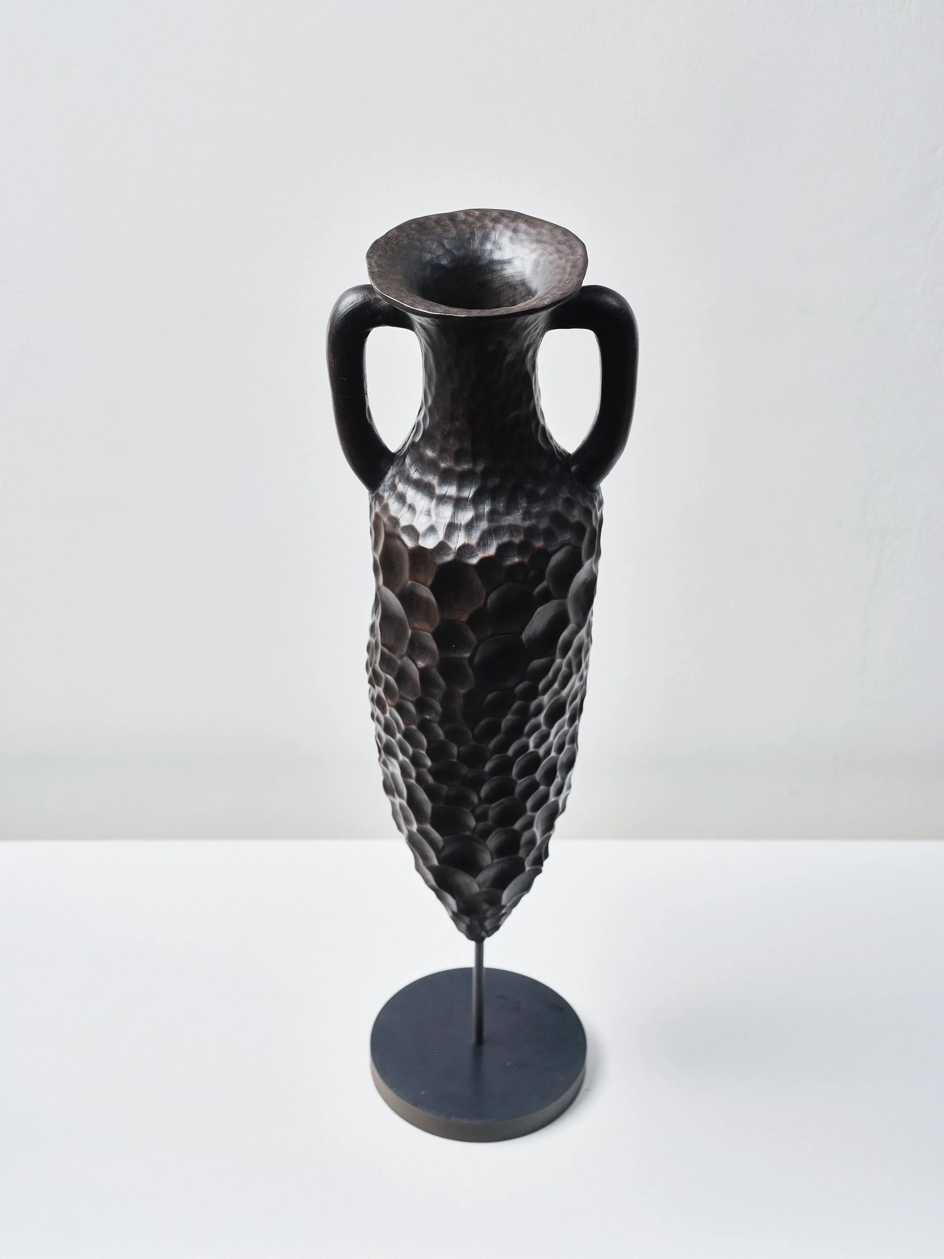 A tall black textured Roman Amphora, hand carved from London Plane with heavy eroded looking honeycomb pattern resembling beaten metal or ceramic, narrow base, two handles, and a flared top, displayed on a pedestal against a white background.