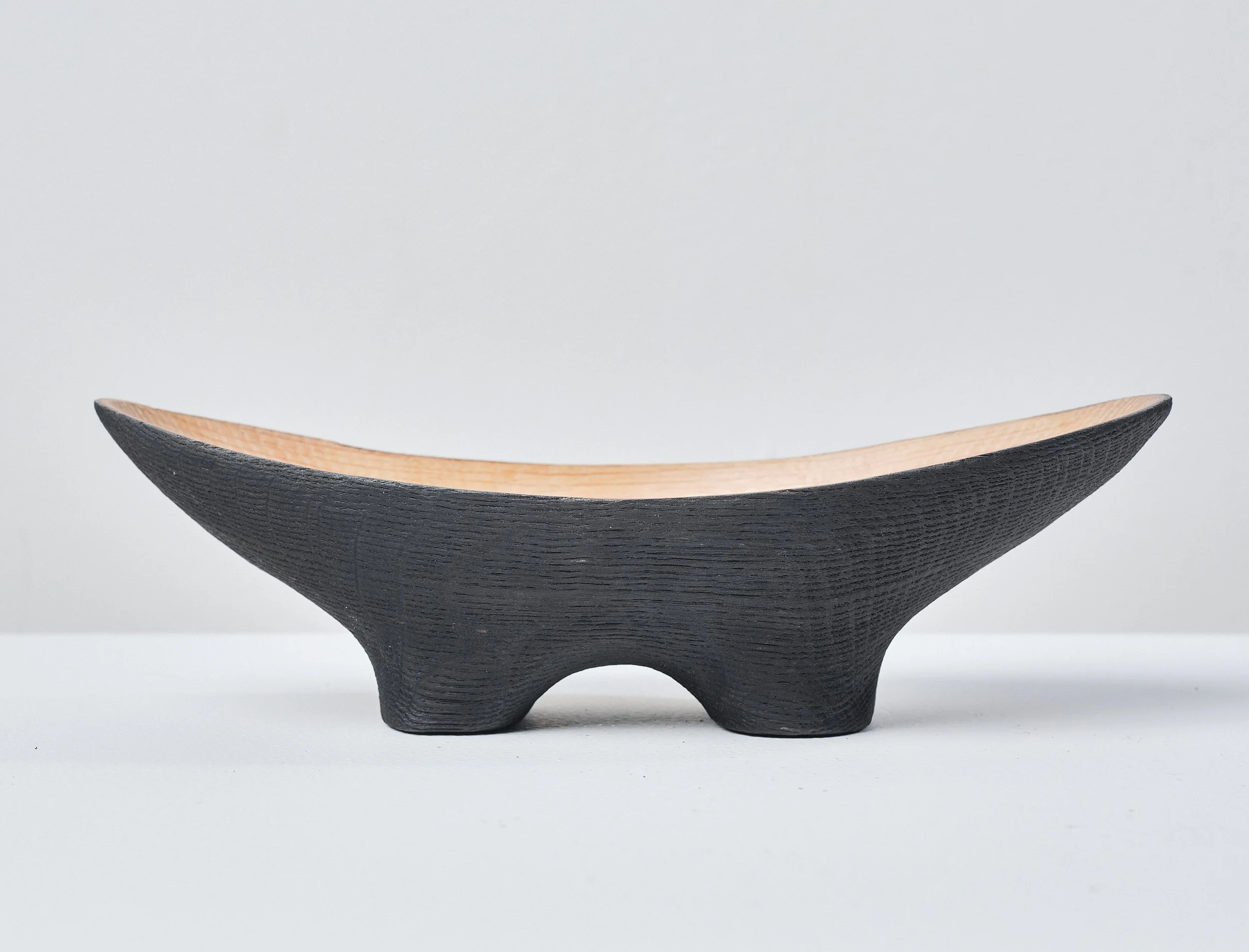 A small wooden bowl made from a salvaged oak beam, with a black exterior and a natural wood interior, placed on a white surface against a plain grey background.