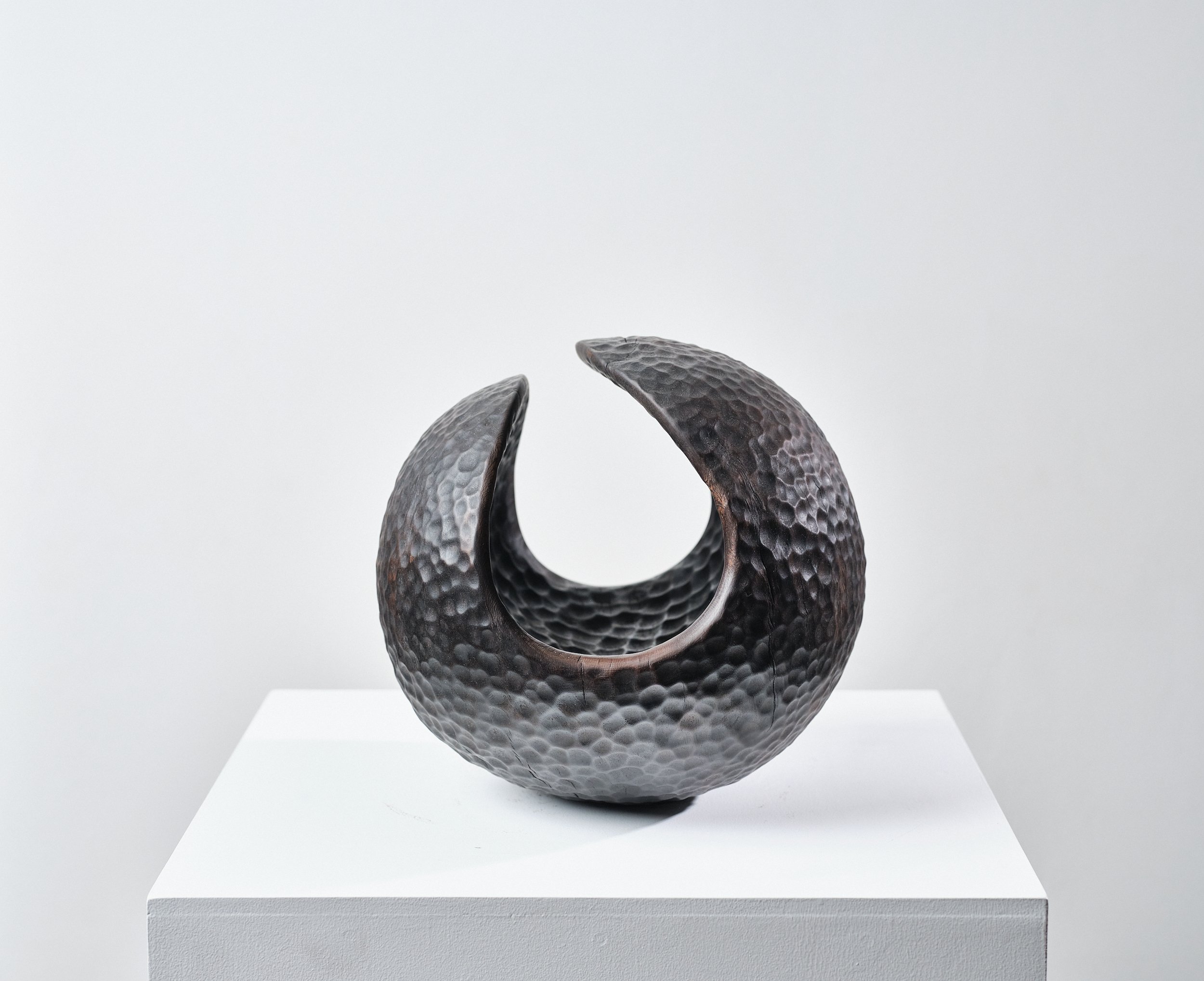 A black, hammered-metal texture sculpture resembling an incomplete circular form with two open, curved ends, displayed on a white pedestal against a plain background in a gallery.