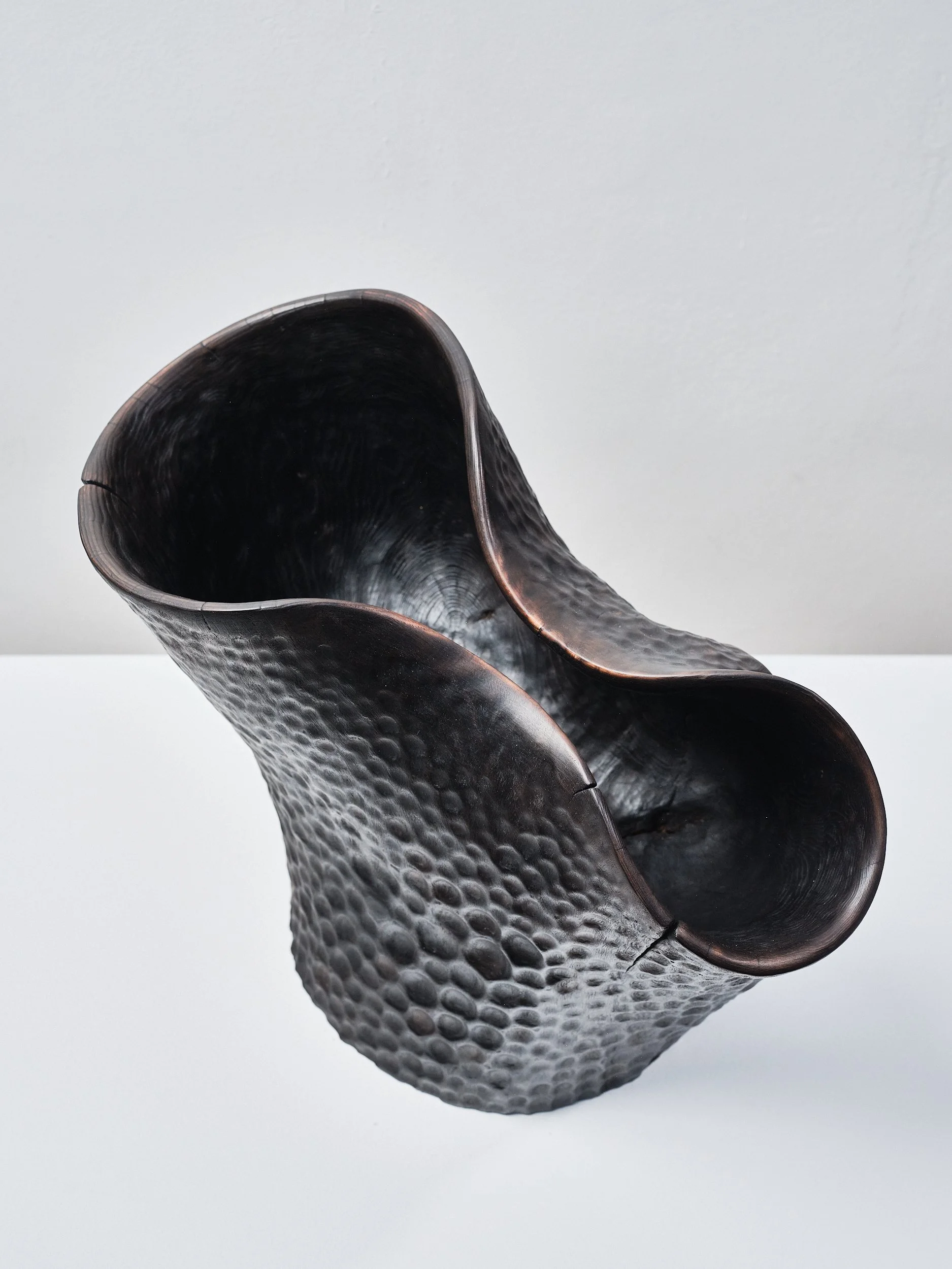 A black textured carved wood functional art piece with an irregular, ruffled opening and hammered metal finish, displayed on a white pedestal against a plain background.