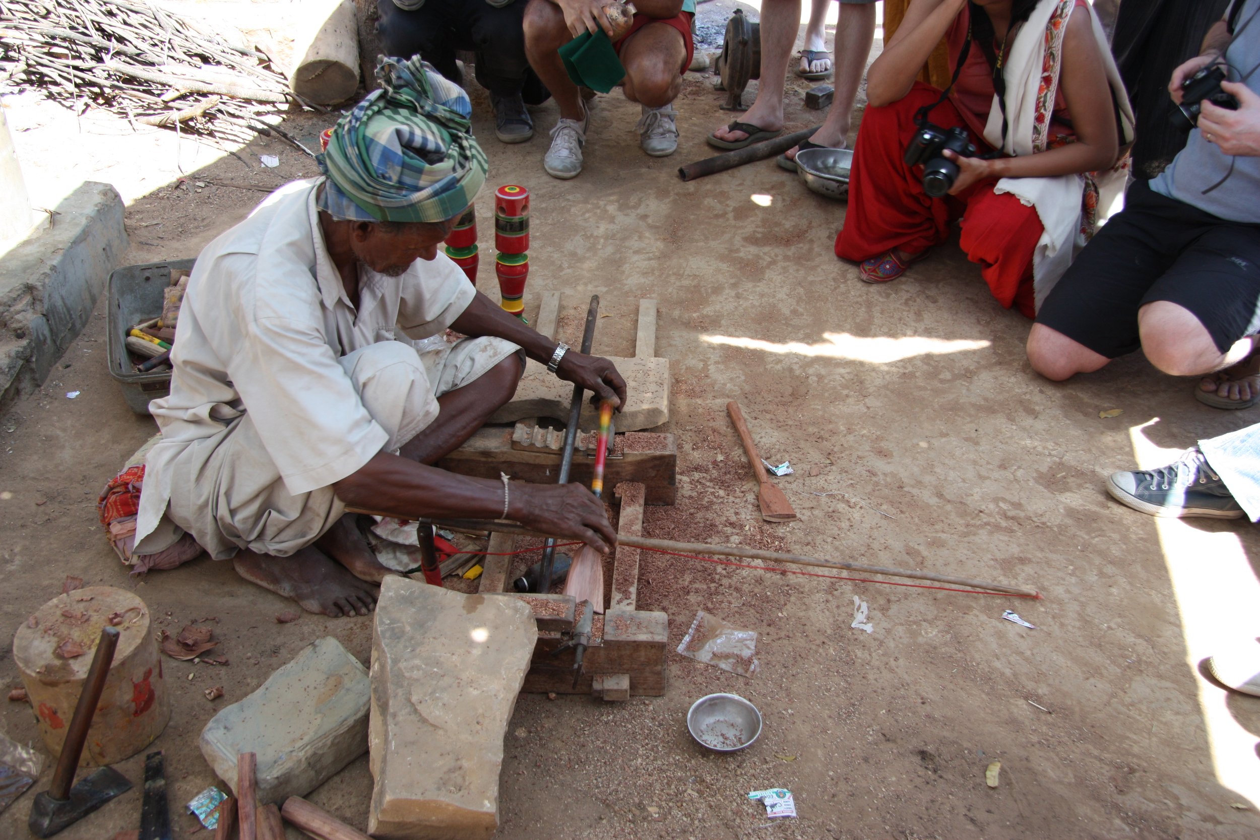 Craftsman Bhavik Bhavchaya is sitting on the ground, working on a lacquer craft, surrounded by spectators from the Royal College of Art with cameras. The setting is an outdoor workshop in india.