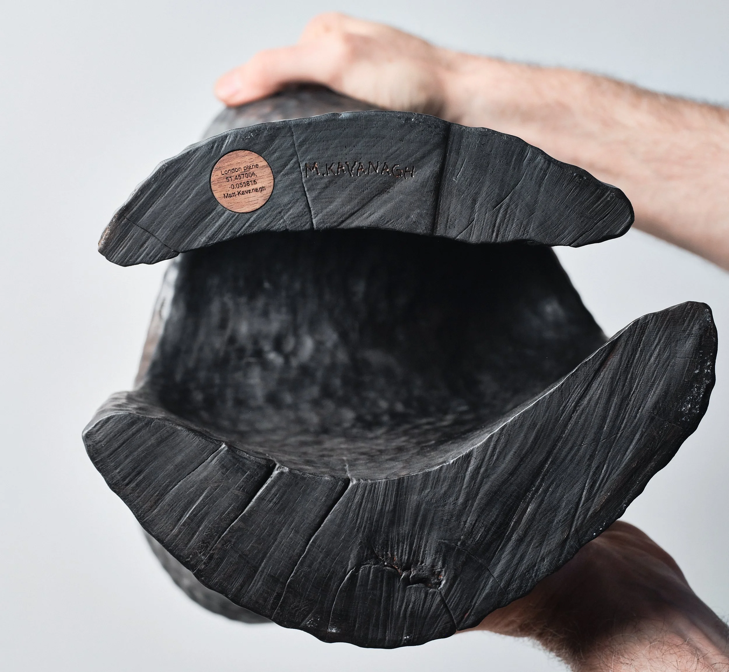 Hands holding a large, contemporary carved wood sculpture, revealing the hollow base and a label engraved with coordinates of where the tree was located to continue its narrative.
