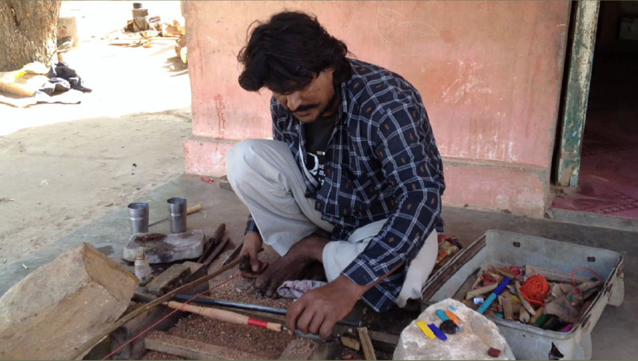 Craftsman Yunas Bhai is sitting on the ground, working on a craft project using tools and materials on a small workspace outside a building. Various tools and crafting supplies are nearby.