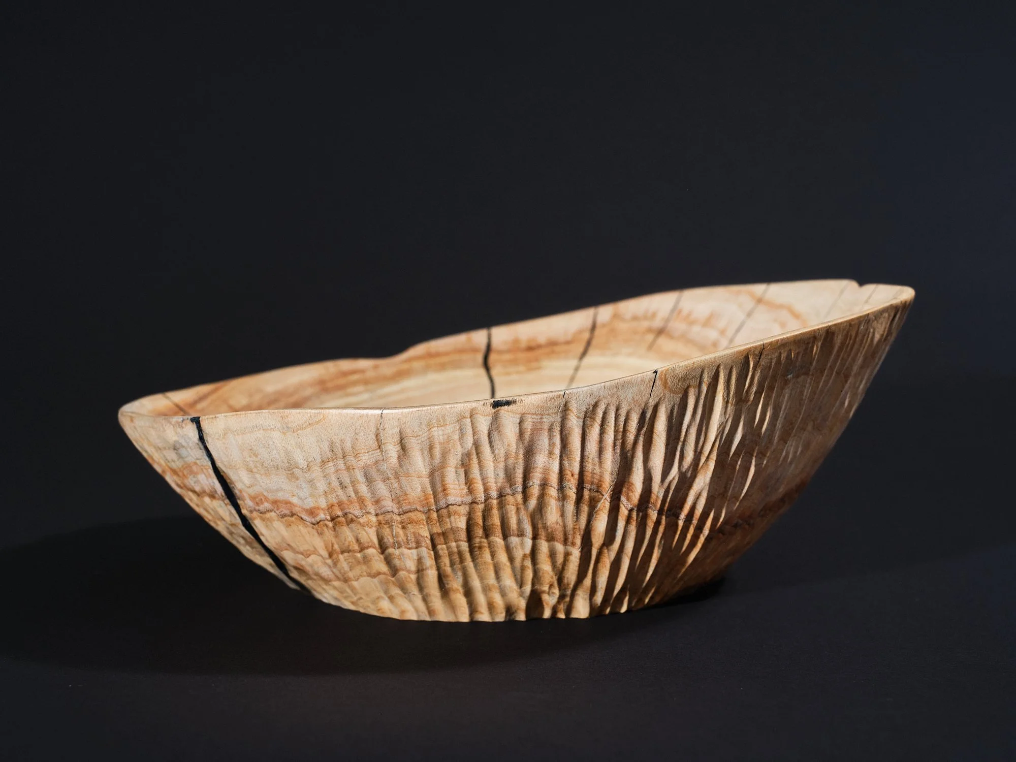 An ash sculpture in a natural waxed and polished wood colour with cracks and splits repaired Kintsugi-style using black pine resin glue, providing visual contrast to the rest of. placed on a white surface against a plain black background.