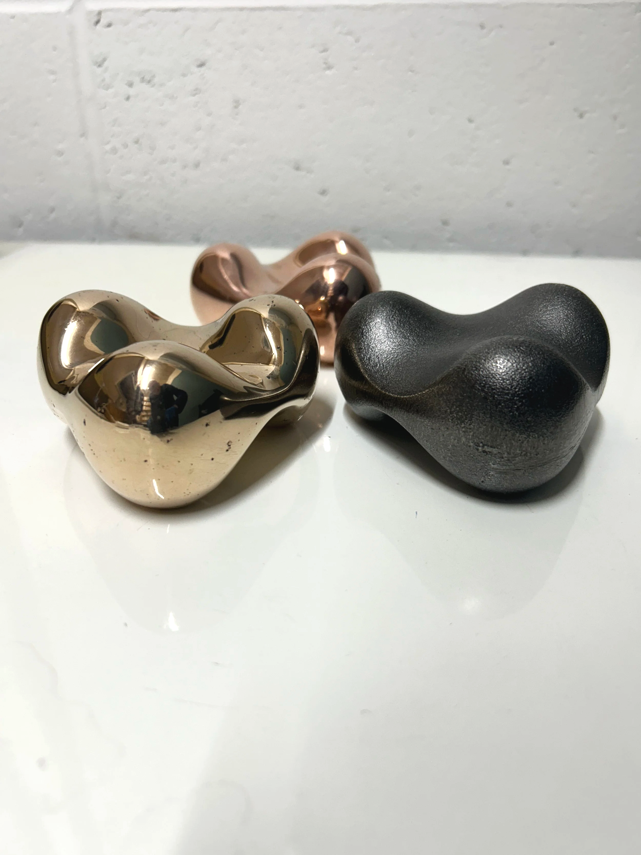 Three abstract, irregularly shaped metallic sculptures in gold, rose gold, and black colours on a white surface with a plain, light-coloured wall in the background.