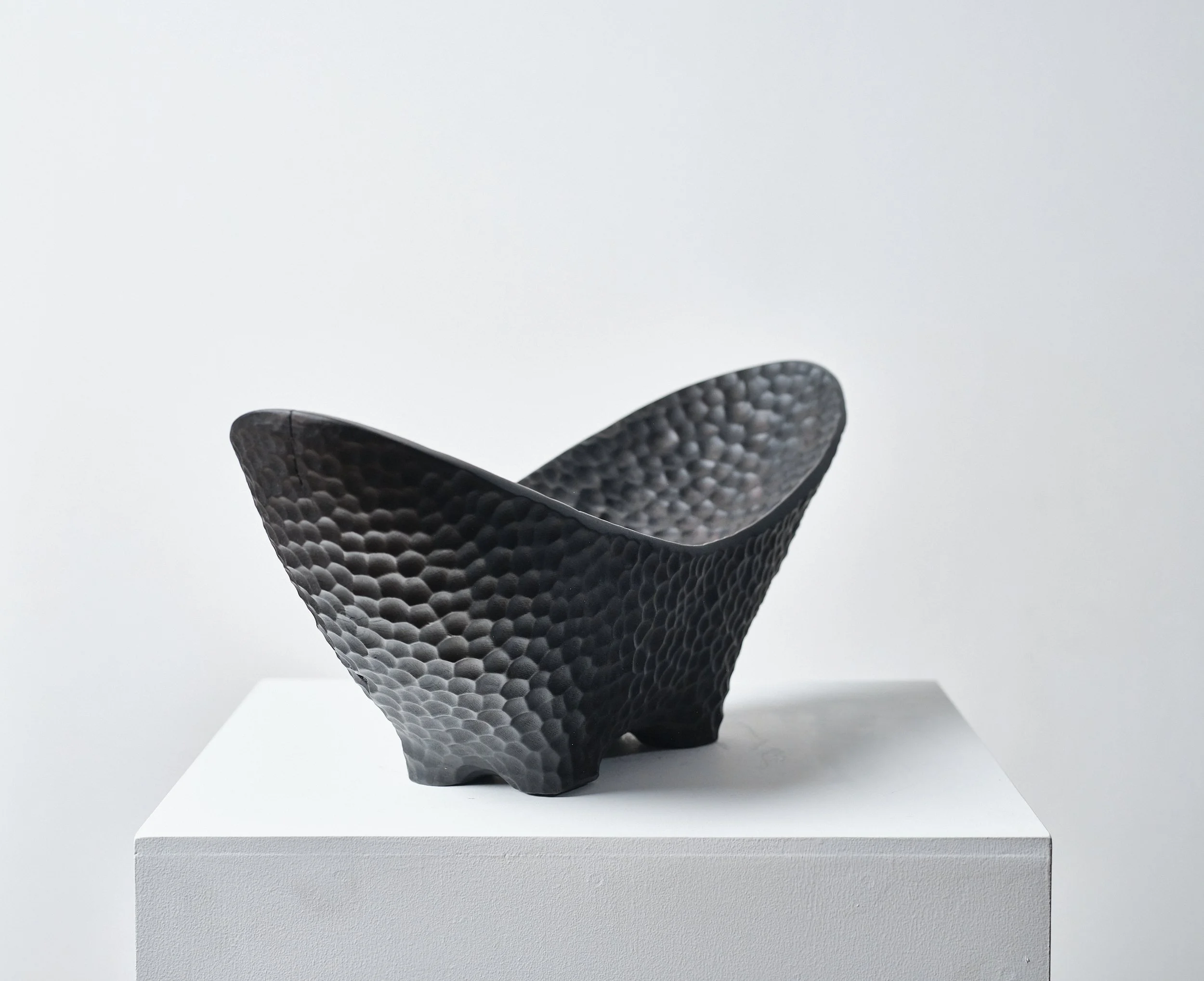 Black textured bowl made from scorched and waxed London Plane timber with an organic shape, displayed on a white pedestal in a gallery setting.