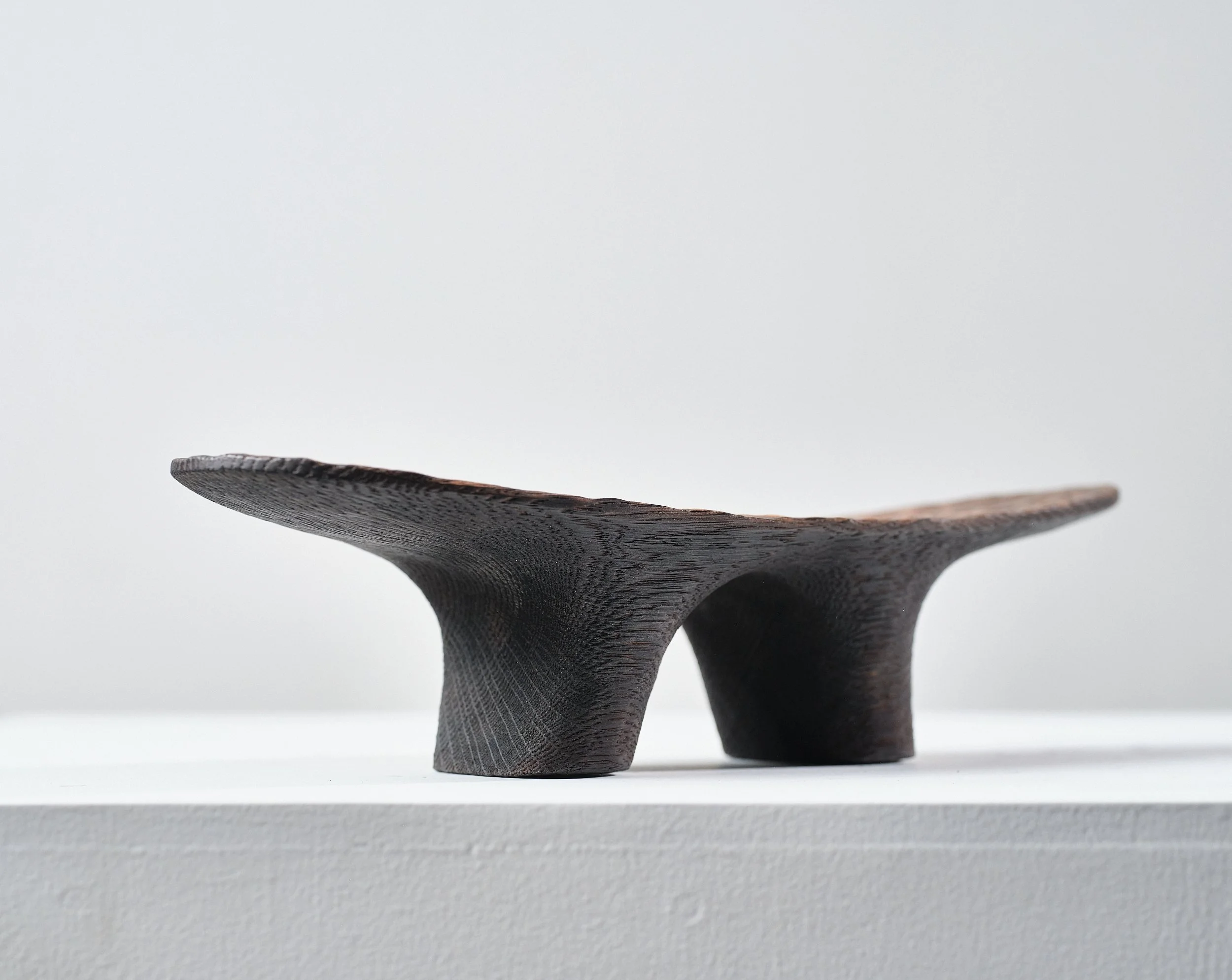 A small, oval-shaped sculpture made from oak from a salvaged beam. The base has two legs and are charred with heavy grain. Sits on a white table against a white wall.