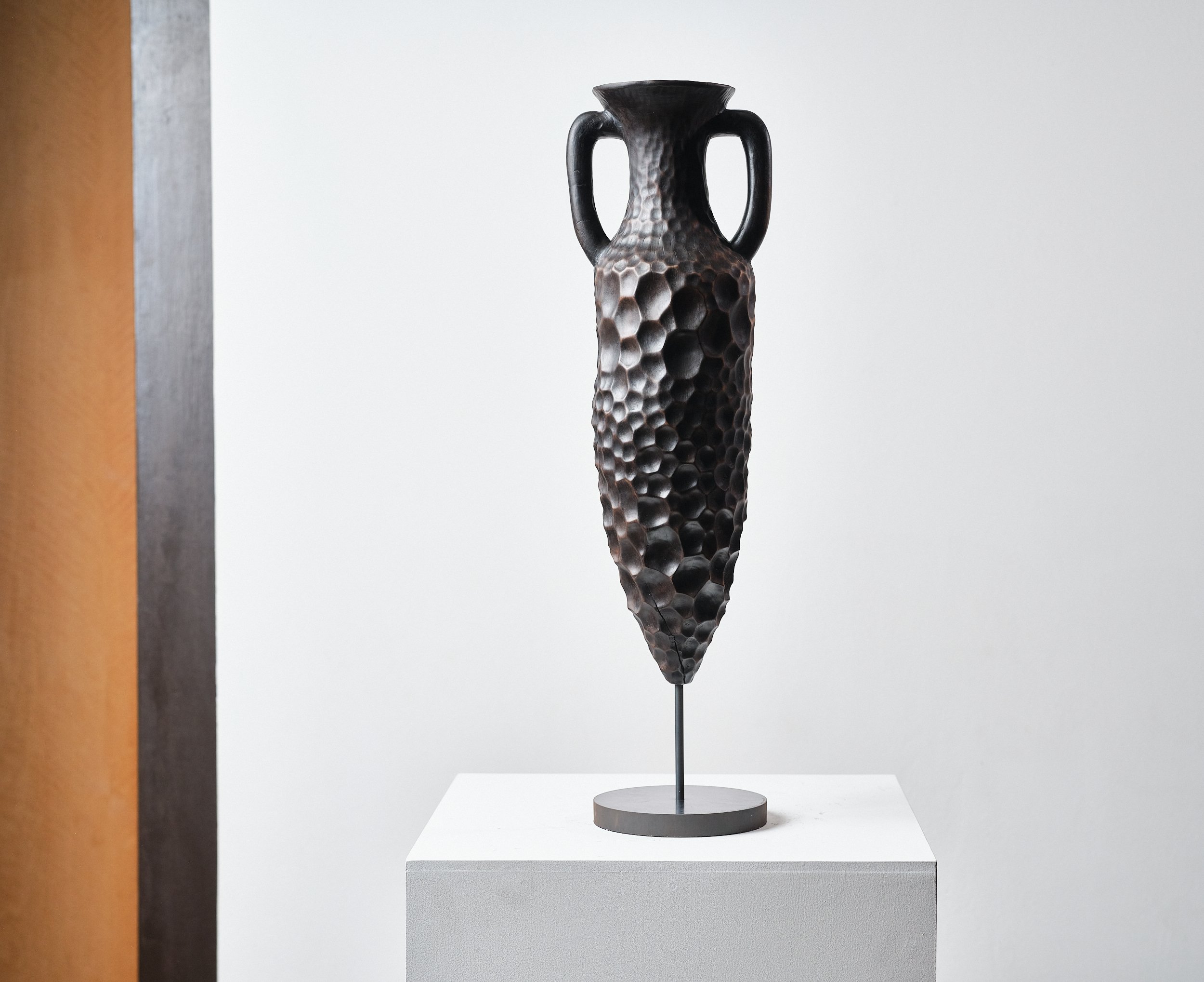 Sculpture of a tall, textured vase, hand carved from London Plane with heavy eroded looking honeycomb pattern resembling beaten metal or ceramic, narrow base, two handles, and a flared top, displayed on a pedestal against a white background.