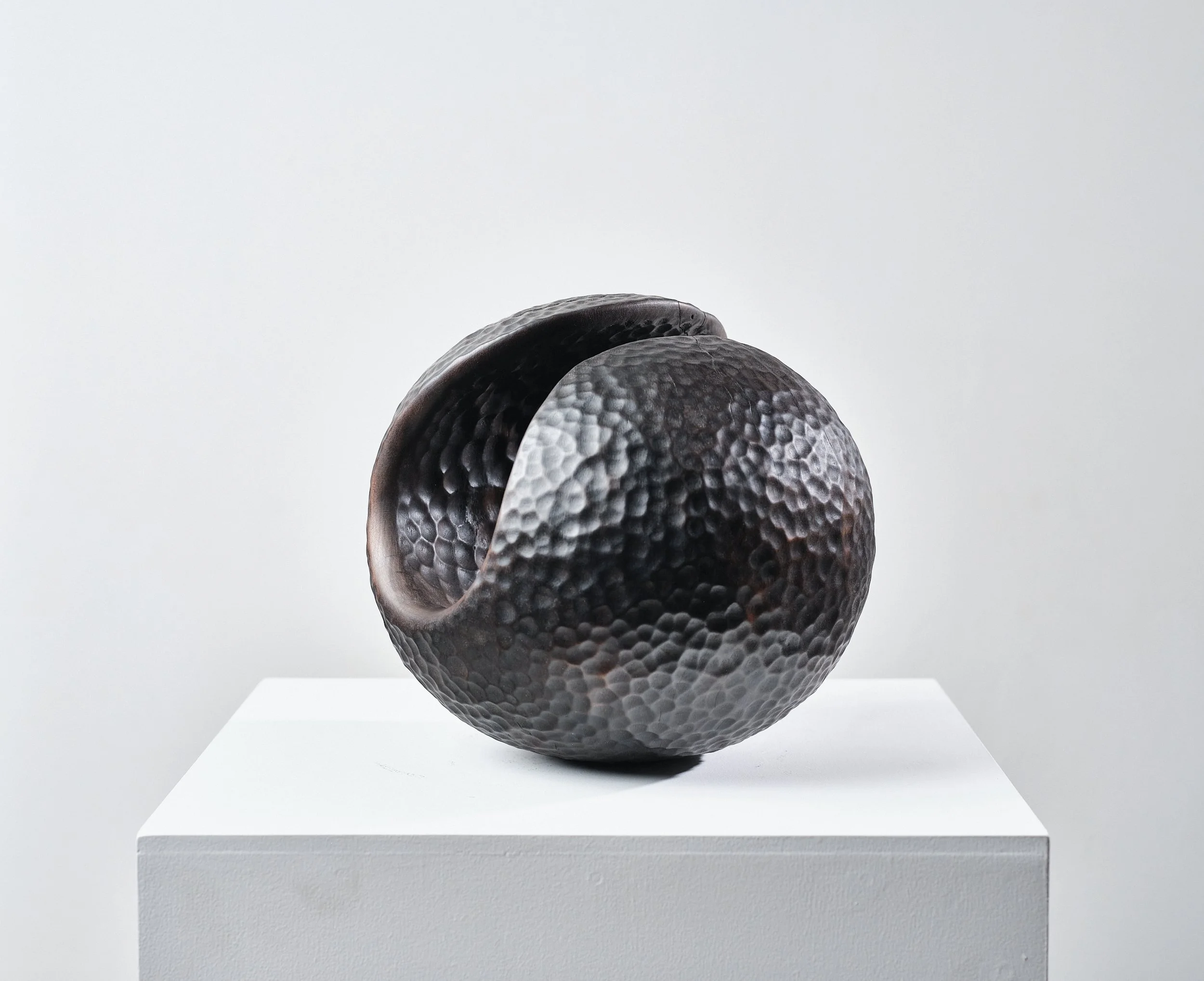 A black textured carved wood spherical sculpture resembling a ceramic vase with an opening in it, displayed on a white pedestal against a plain background in an art gallery.