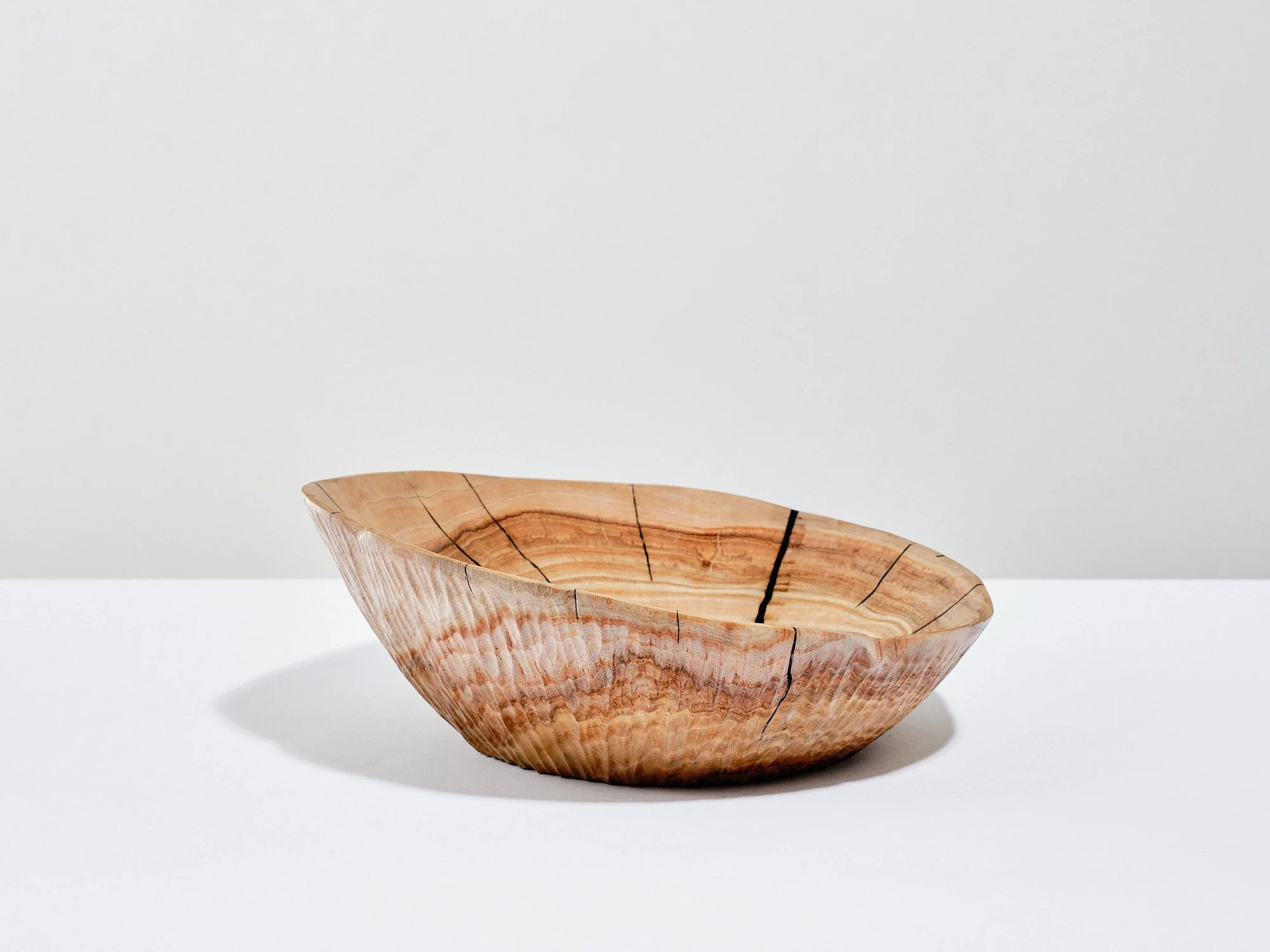 An ash sculpture in a natural waxed and polished wood colour with cracks and splits repaired Kintsugi-style using black pine resin glue, providing visual contrast to the rest of. placed on a white surface against a plain white background.