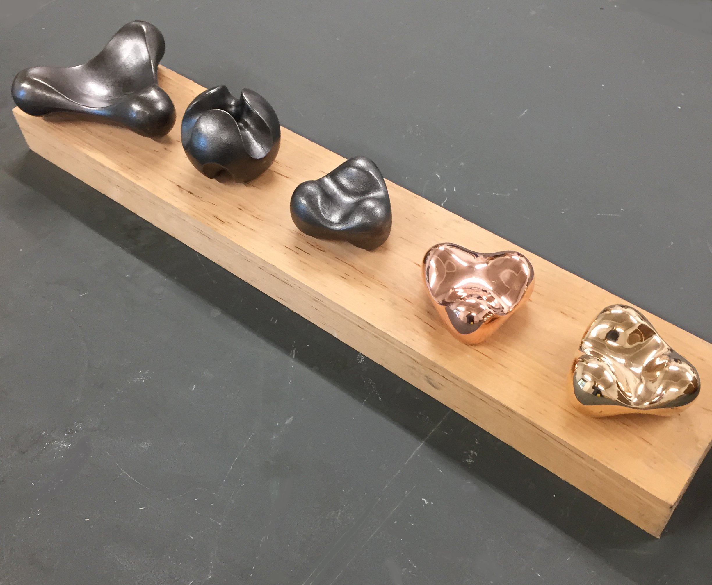 Six metallic functional sculptures in various shapes and colours on a wooden plinth. Made from blackened iron, polished bronze and copper