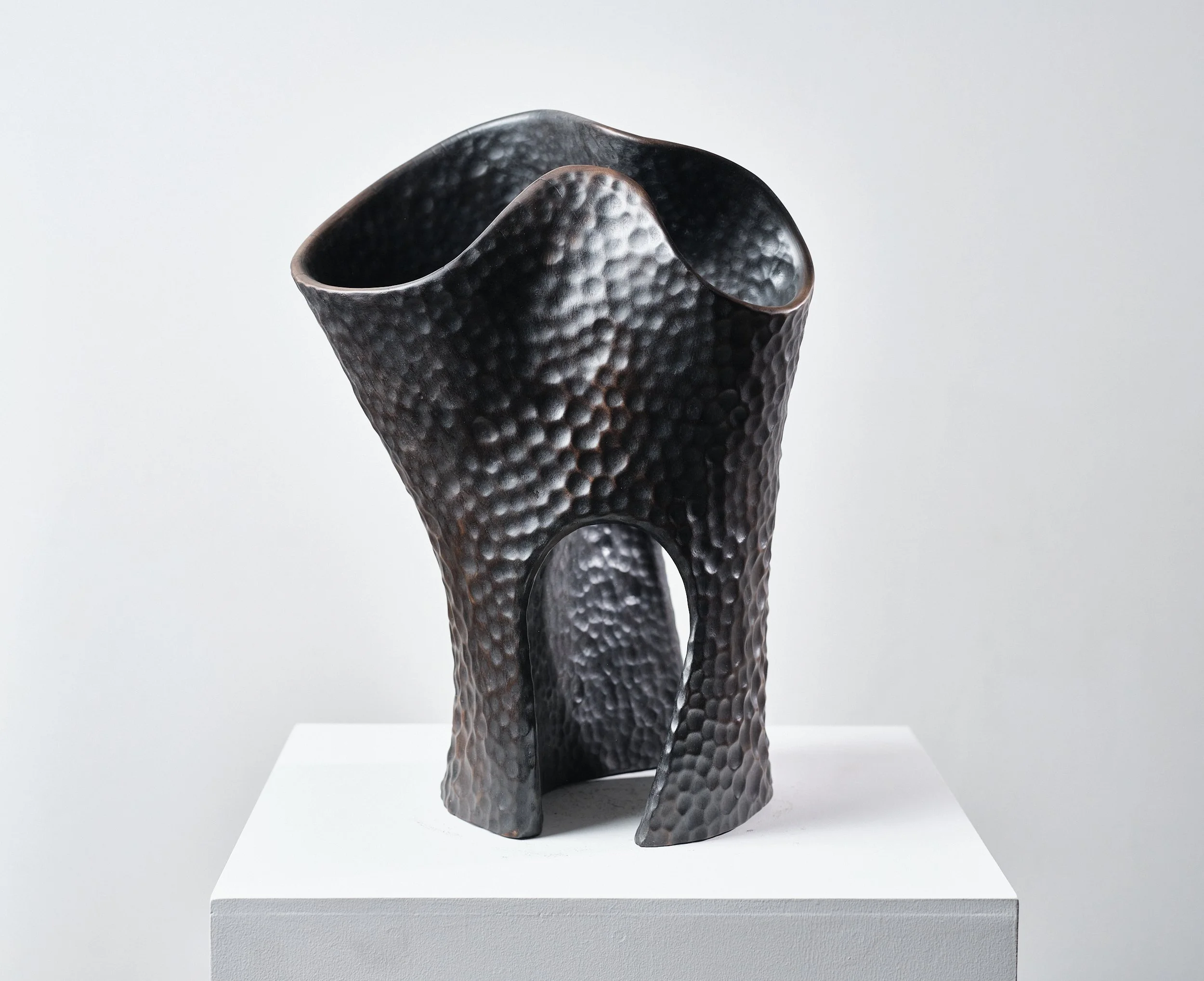Front view of a carved timber functional sculpture with curved opening at the top and hollow base, surface like beaten blackened bronze on white pedestal.