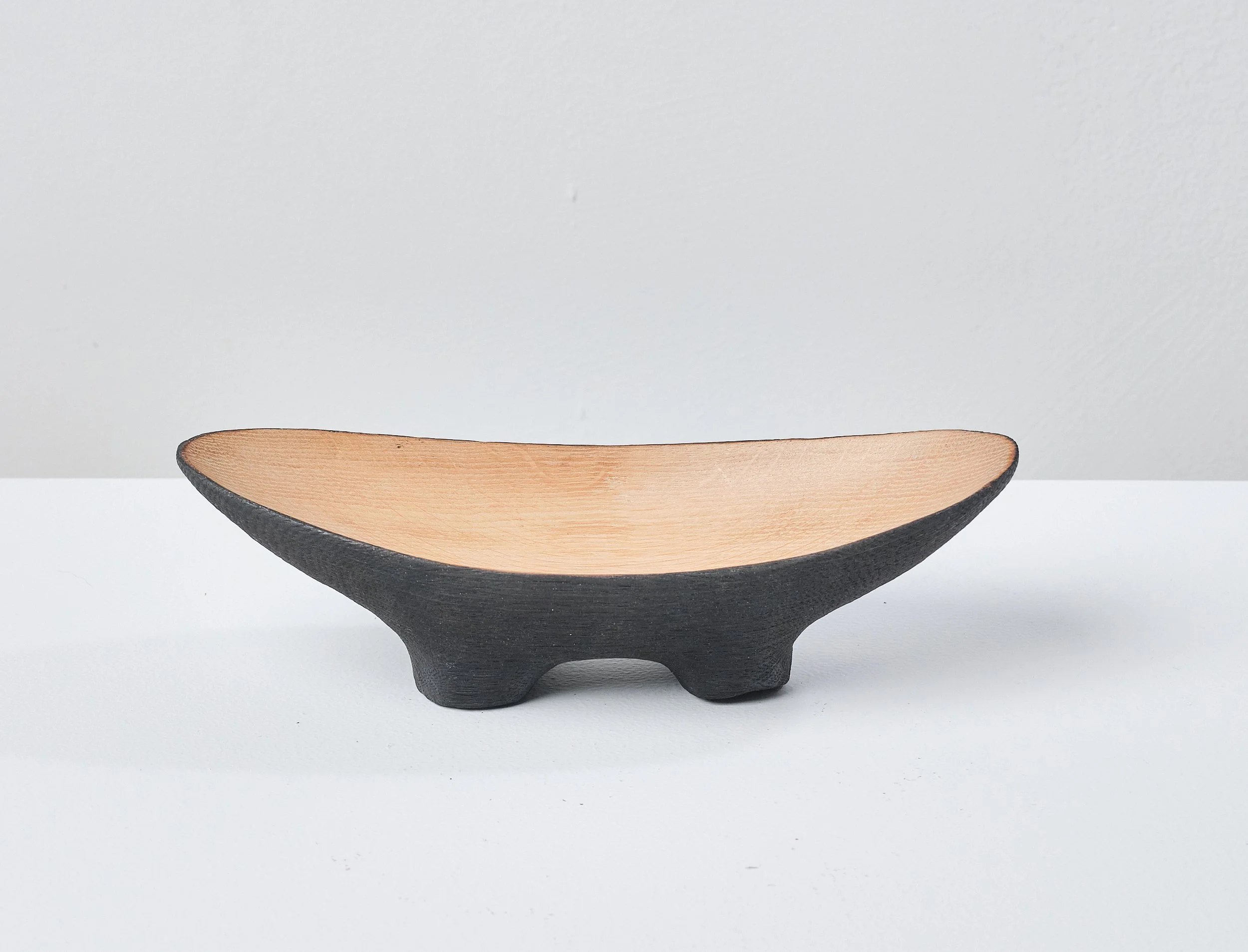A small wooden bowl made from a salvaged oak beam. The inside has a natural oak colour and a waxed sheen. The base is black from scorching and oil finished.  Placed on a white surface against a pale grey wall