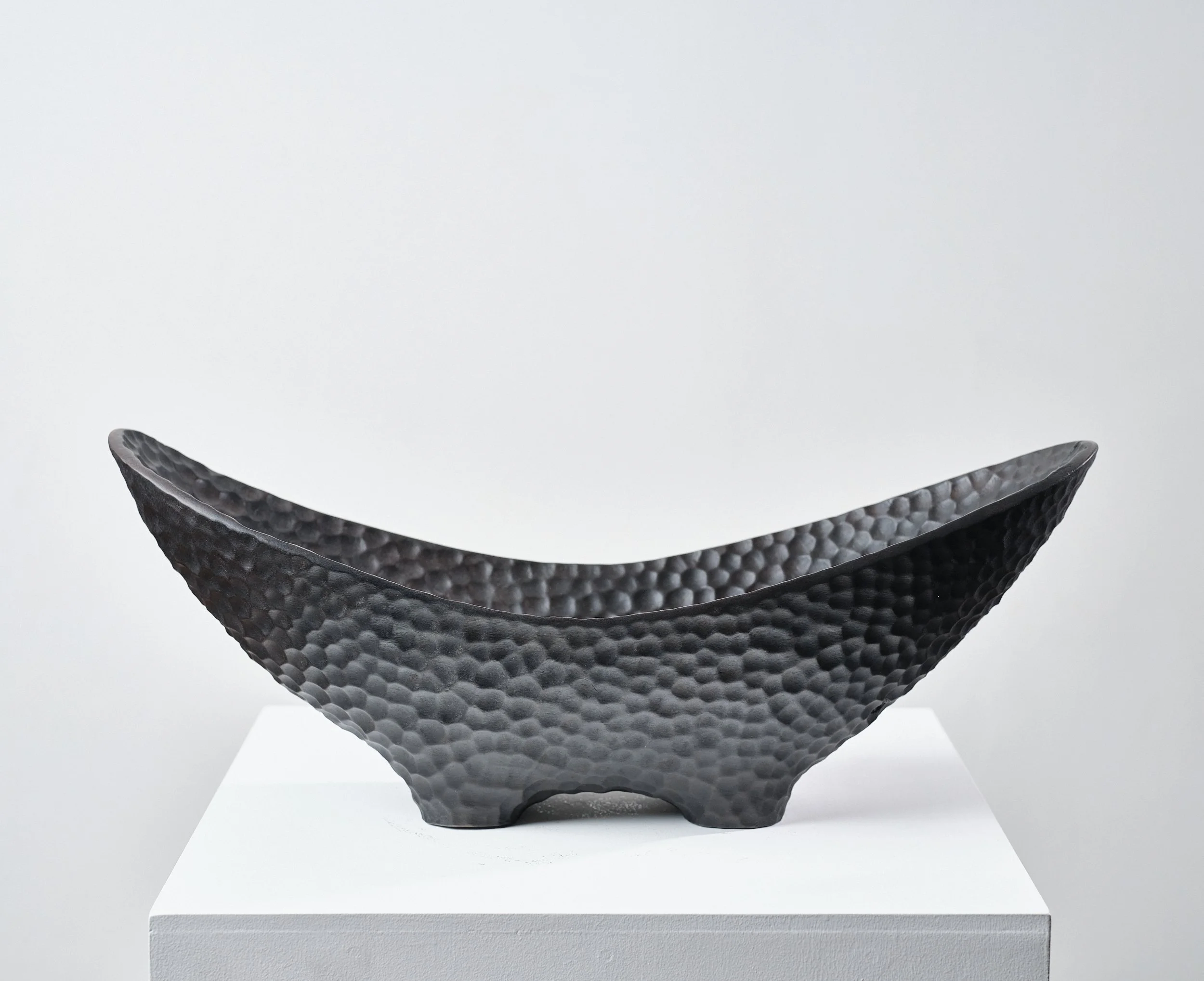 Black textured bowl made from scorched and waxed London Plane timber with an organic shape, displayed on a white pedestal in a gallery setting.