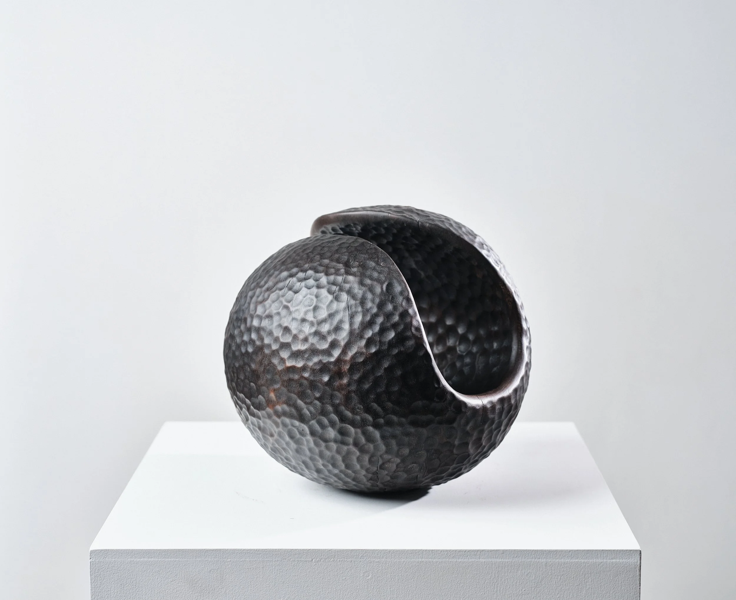 A black textured carved wood spherical sculpture resembling a beaten metal surface with an opening in it, displayed on a white pedestal against a plain background in an art gallery.