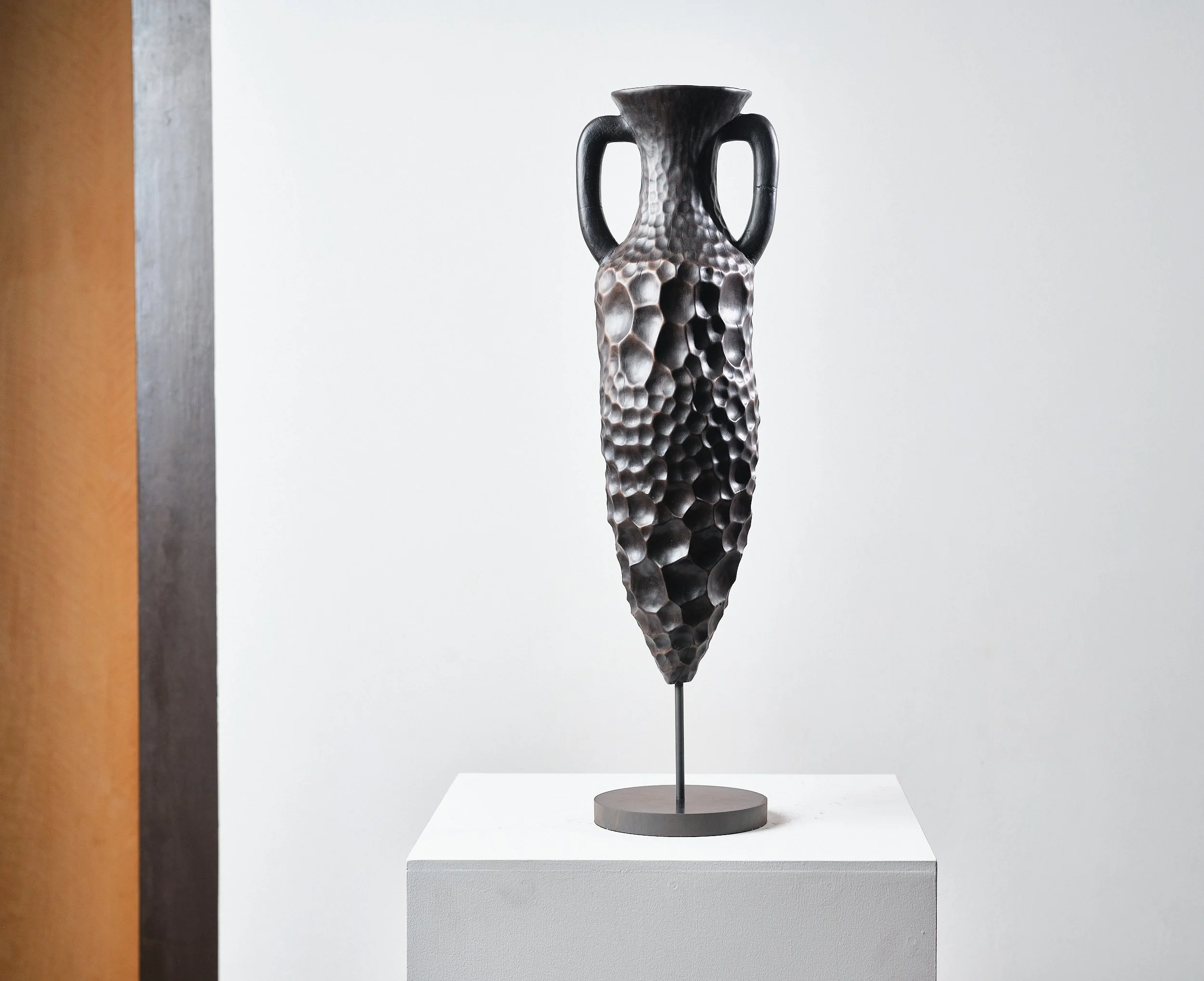 Sculpture of a tall, textured vase, hand carved from London Plane with heavy eroded looking honeycomb pattern resembling beaten metal or ceramic, narrow base, two handles, and a flared top, displayed on a pedestal against a white background.
