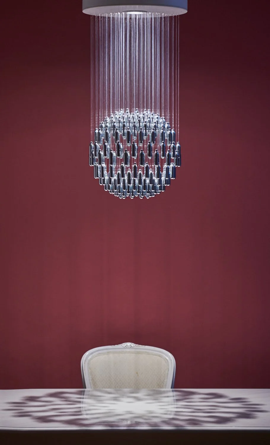 NOS Sphere 228 chandelier, made from salvaged nitrous oxide canisters, suspended over a white table and chair with a pattern of light on the reflective table top. Against a maroon wall.