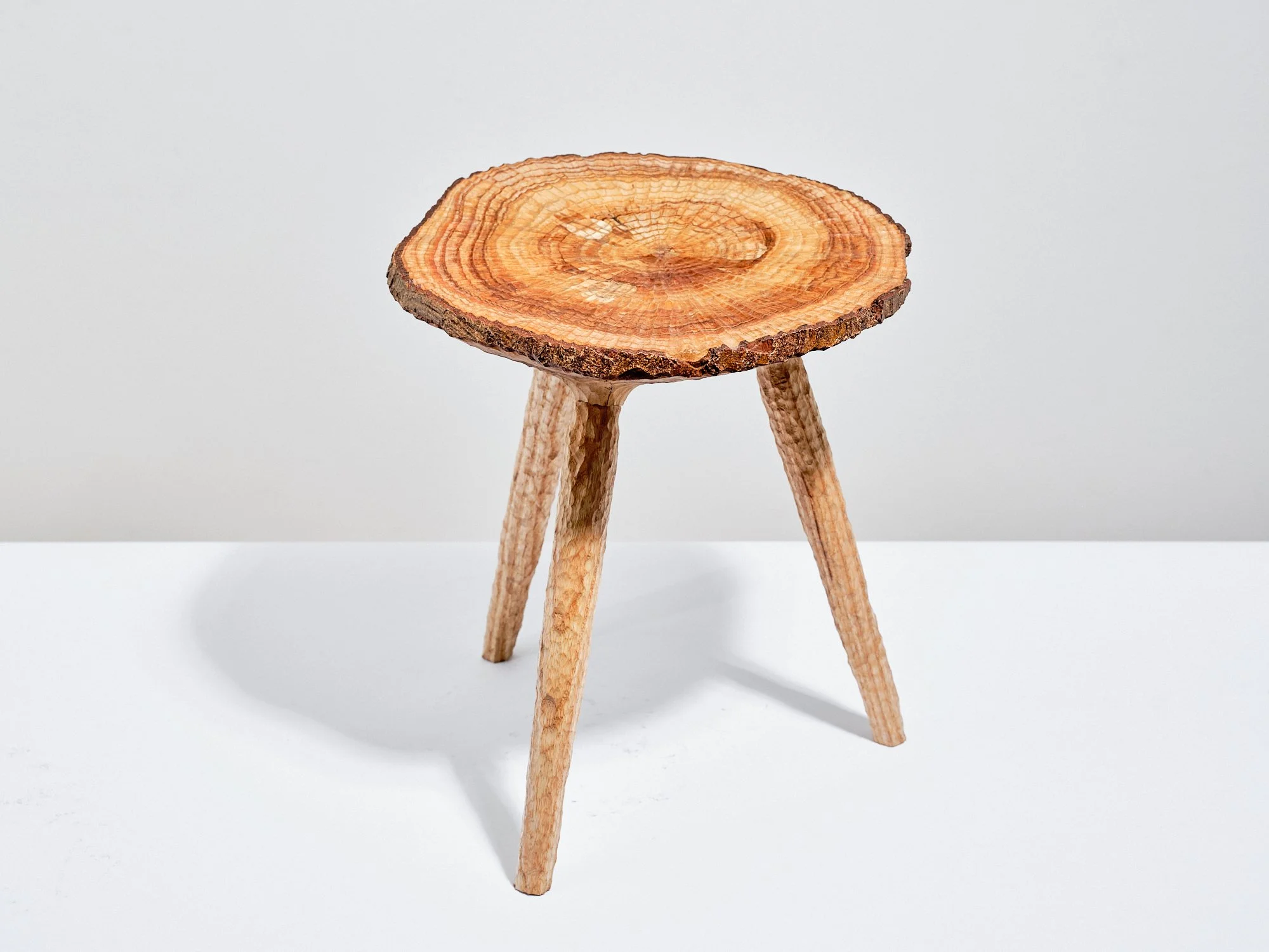 A small free carved, contemporary crafted, wooden milking stool made from a disc of ash with edge grain and three angled ash legs, oil finished, set against a white background.