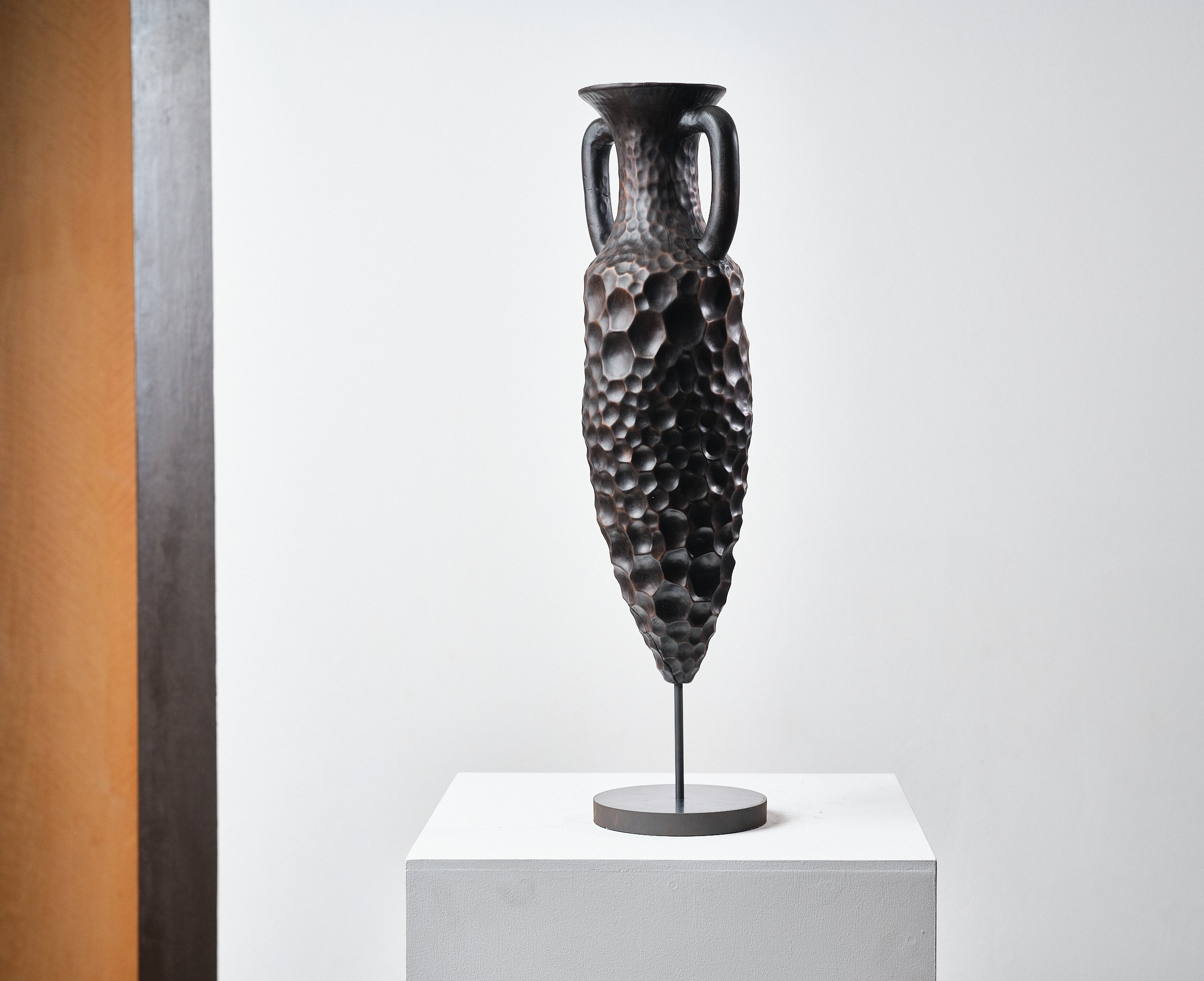 Black sculpture of a tall, elongated vase with a heavy honeycomb textured pattern, mounted on a round black metal base, displayed on a white pedestal in a gallery.