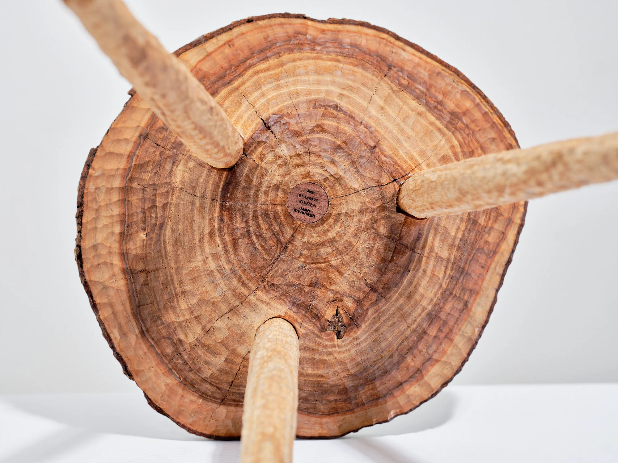 Underside view of free carved, wooden milking stool made from a disc of ash with edge grain and three angled ash legs, oil finished. In the centre there is an engraved badge with coordinates of the place the tree grew, set against a white background.