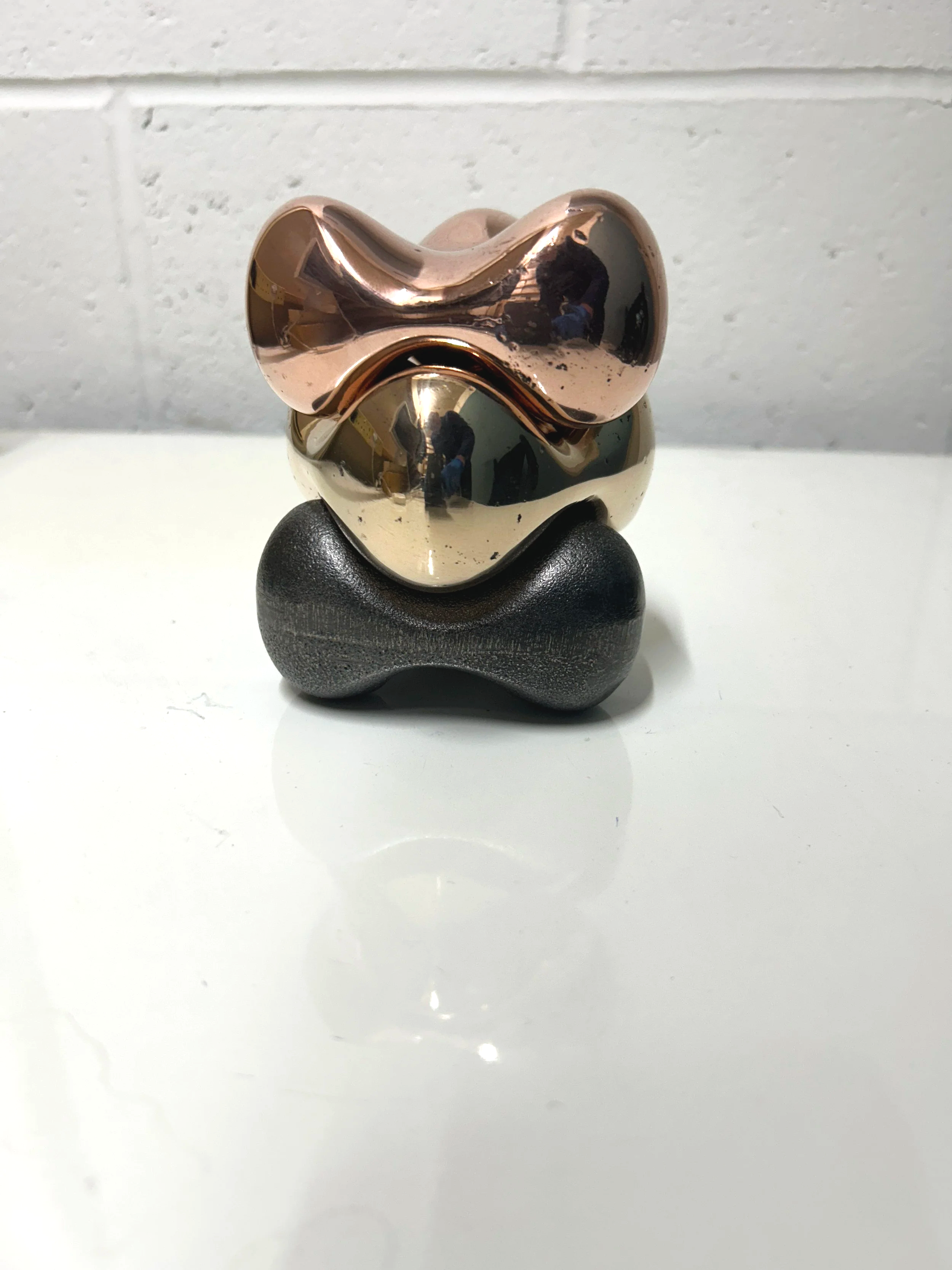 Stack of three small abstract sculptures in different metallic finishes, including rose gold, gold, and black, on a white surface against a white brick wall.