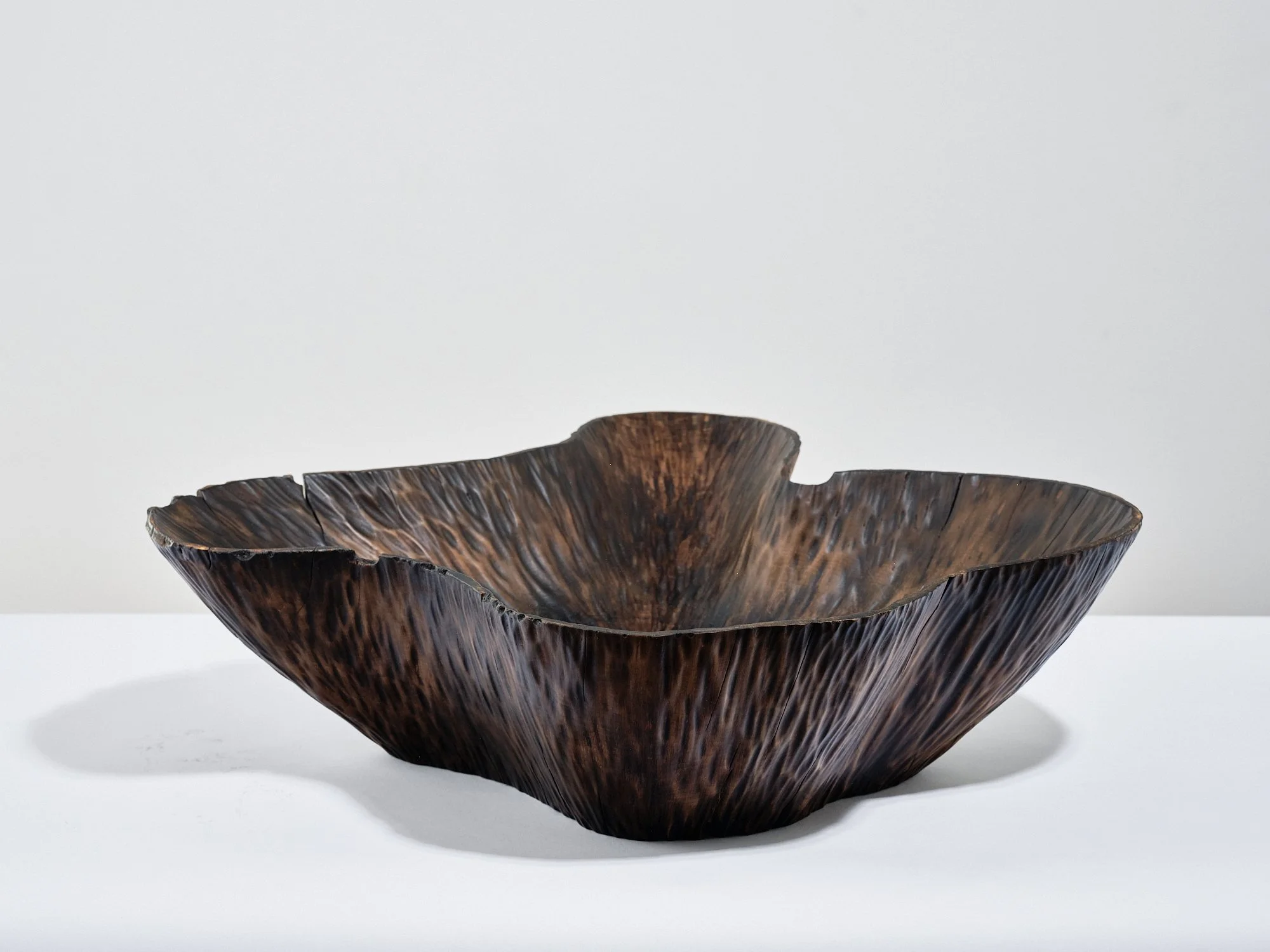 A wooden bowl free-carved from Horse Chestnut scorched and sanded to provide a conker brown finish and a textured irregular surface, placed on a white table against a plain light background.