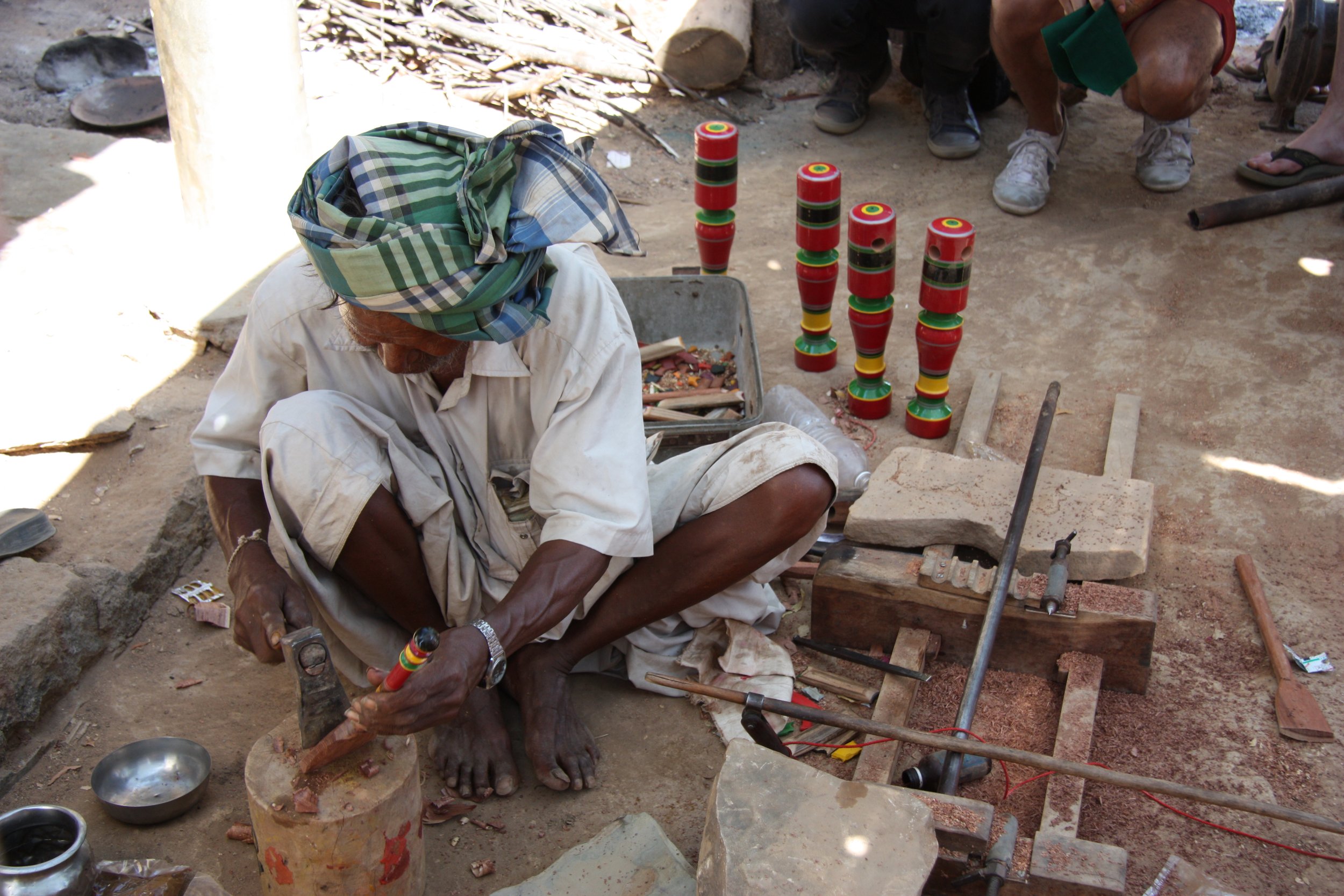 A man wearing a plaid turban and white clothes is sitting cross-legged on the ground, working on a small craft project with a hammer. Surrounding him are tools, stones, a box of colourful small objects, and several tall, painted, cylindrical objects.