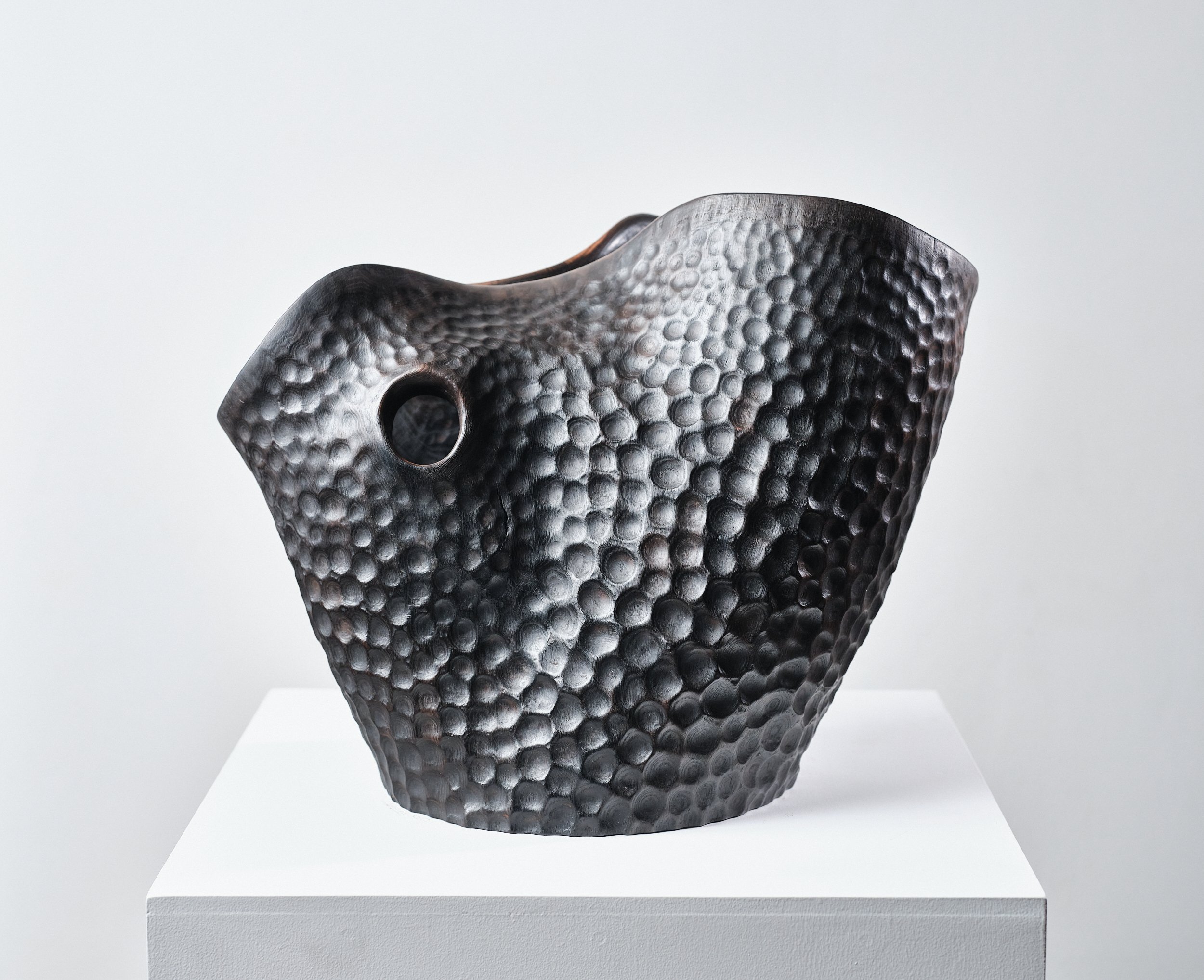 A dark, textured vase carved from green ash, with a hammered & polished pattern, displayed on a white pedestal against a plain grey background.