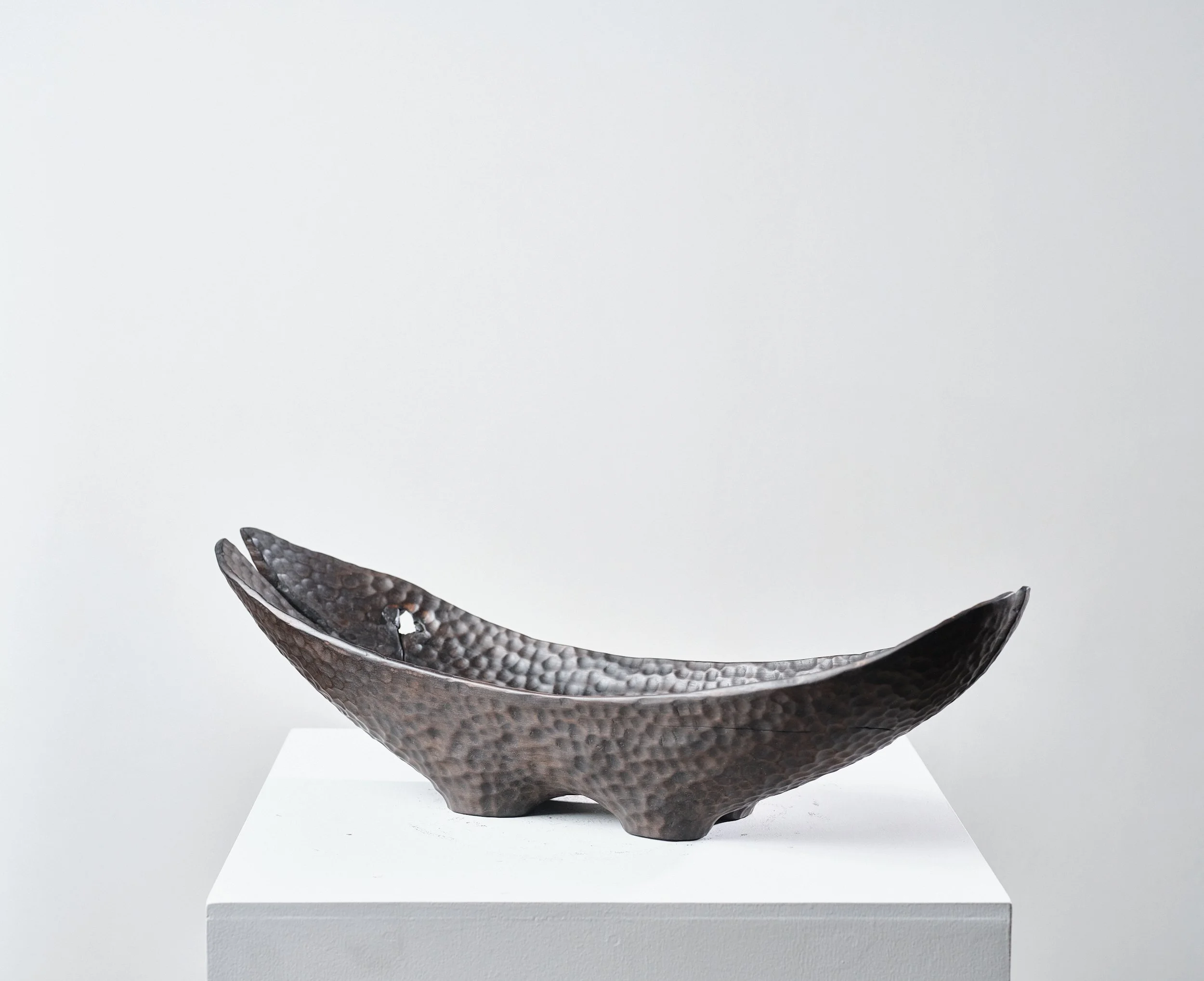 Norwegian Maple Long Bowl with dimpled polished surface that resemble black hammered metal with splits from the timber and a hole on the top, placed on a white plinth against plain white wall.