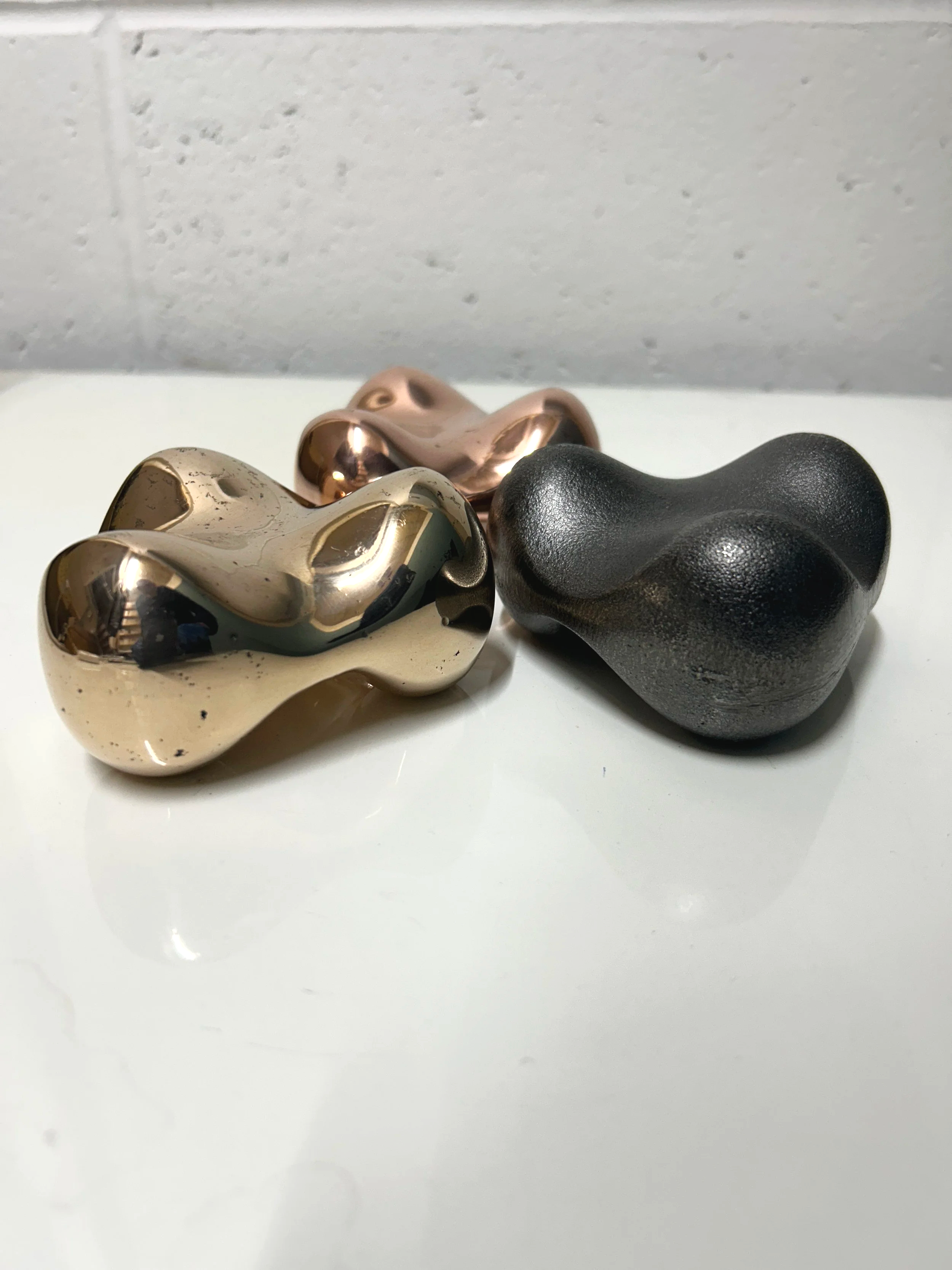 Three abstract, irregularly shaped metallic sculptures in gold, rose gold, and black colours on a white surface with a plain, light-coloured wall in the background.