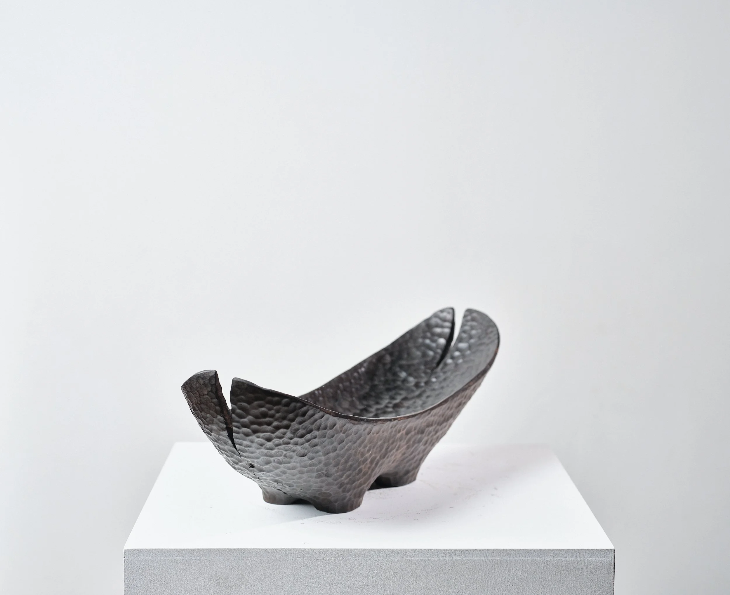 Norwegian Maple Long Bowl with dimpled polished surface that resemble black hammered metal with splits from the timber and a hole on the top, placed on a white plinth against plain white wall.