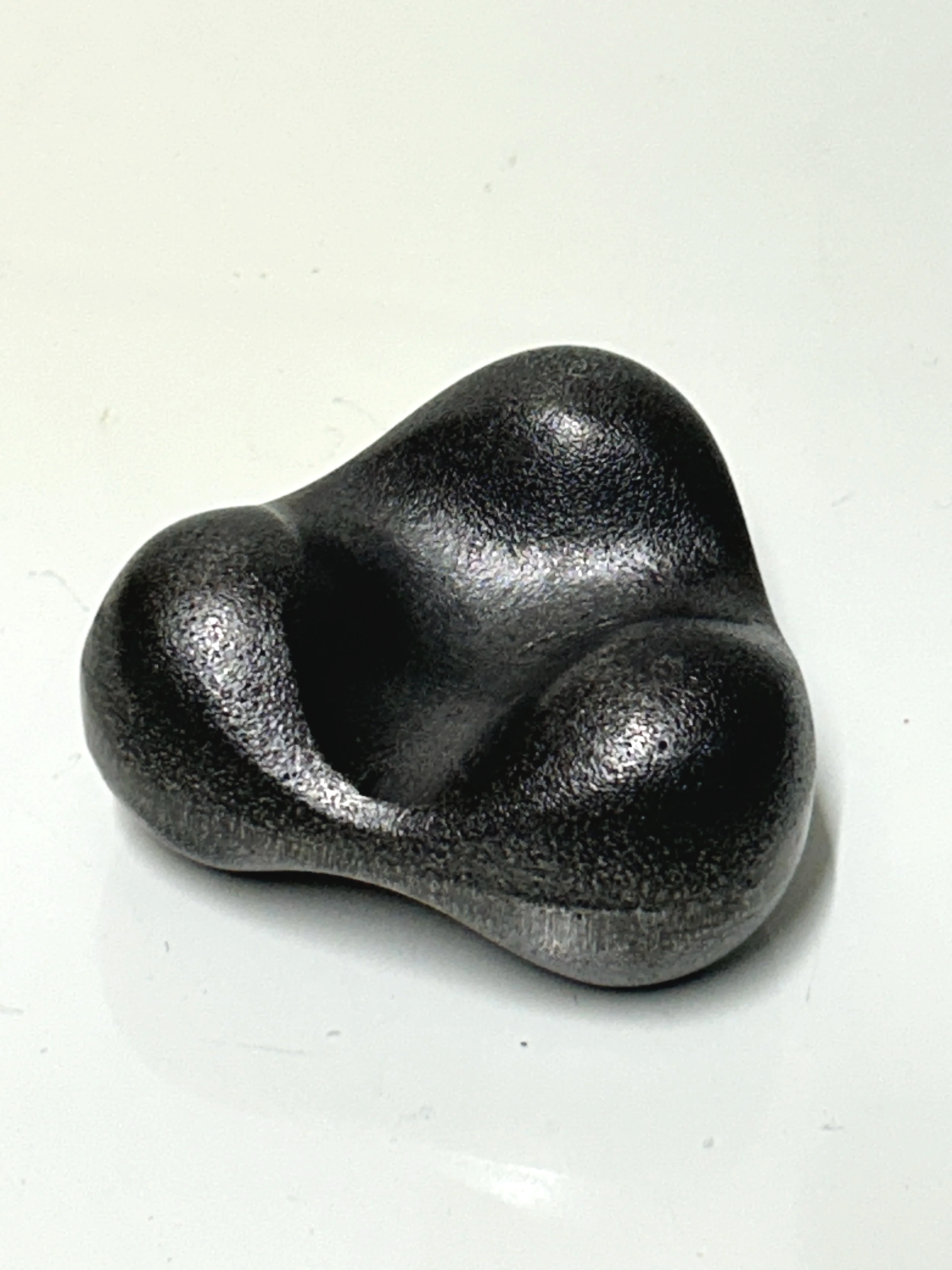 A black, textured, abstract-shaped sculpture with smooth, rounded contours on a white surface.