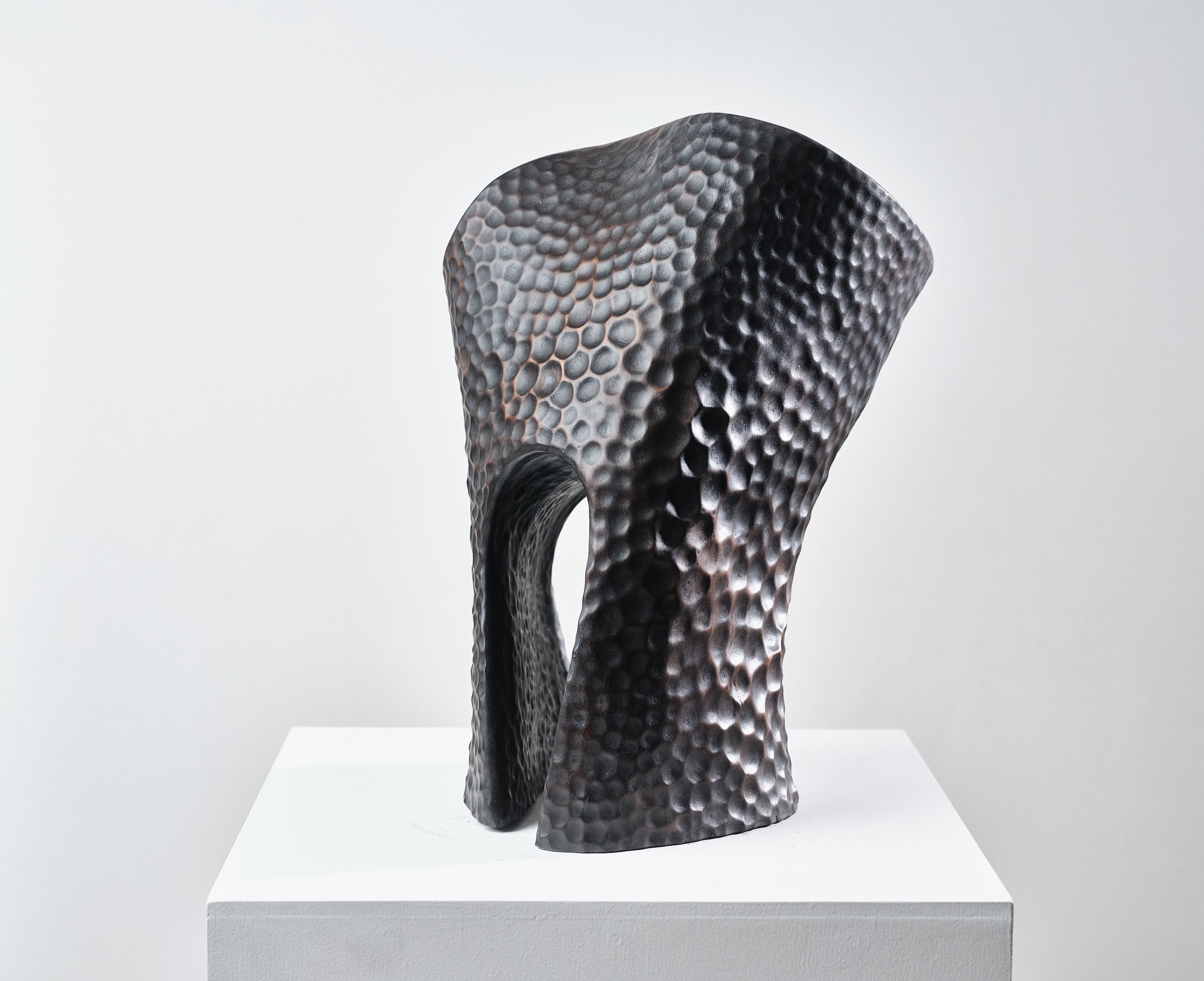 A modern, abstract sculpture with a hammered texture, featuring a curved, hollow shape, displayed on a white pedestal against a plain background.