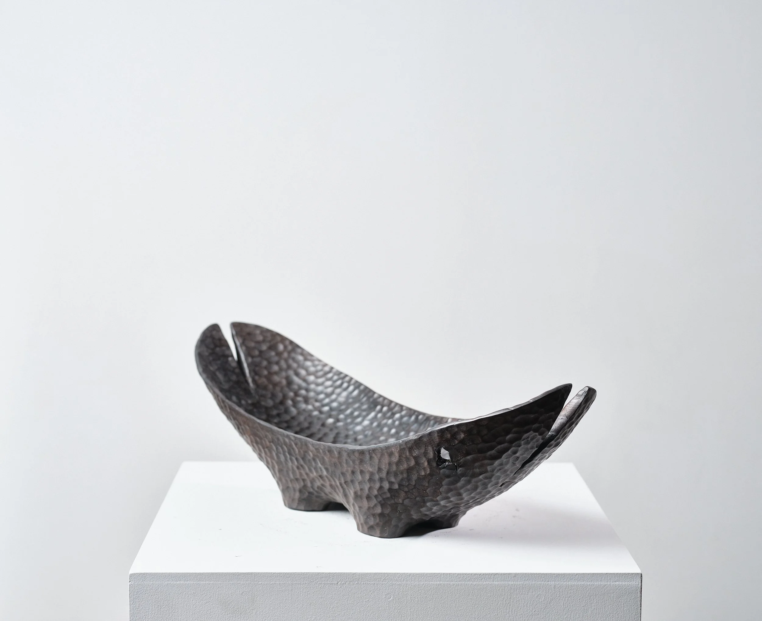Norwegian Maple Long Bowl organic form sitting on small legs. Has a dimpled polished surface that resemble black hammered metal with splits from the timber and a hole on the top, placed on a white plinth against plain white wall.