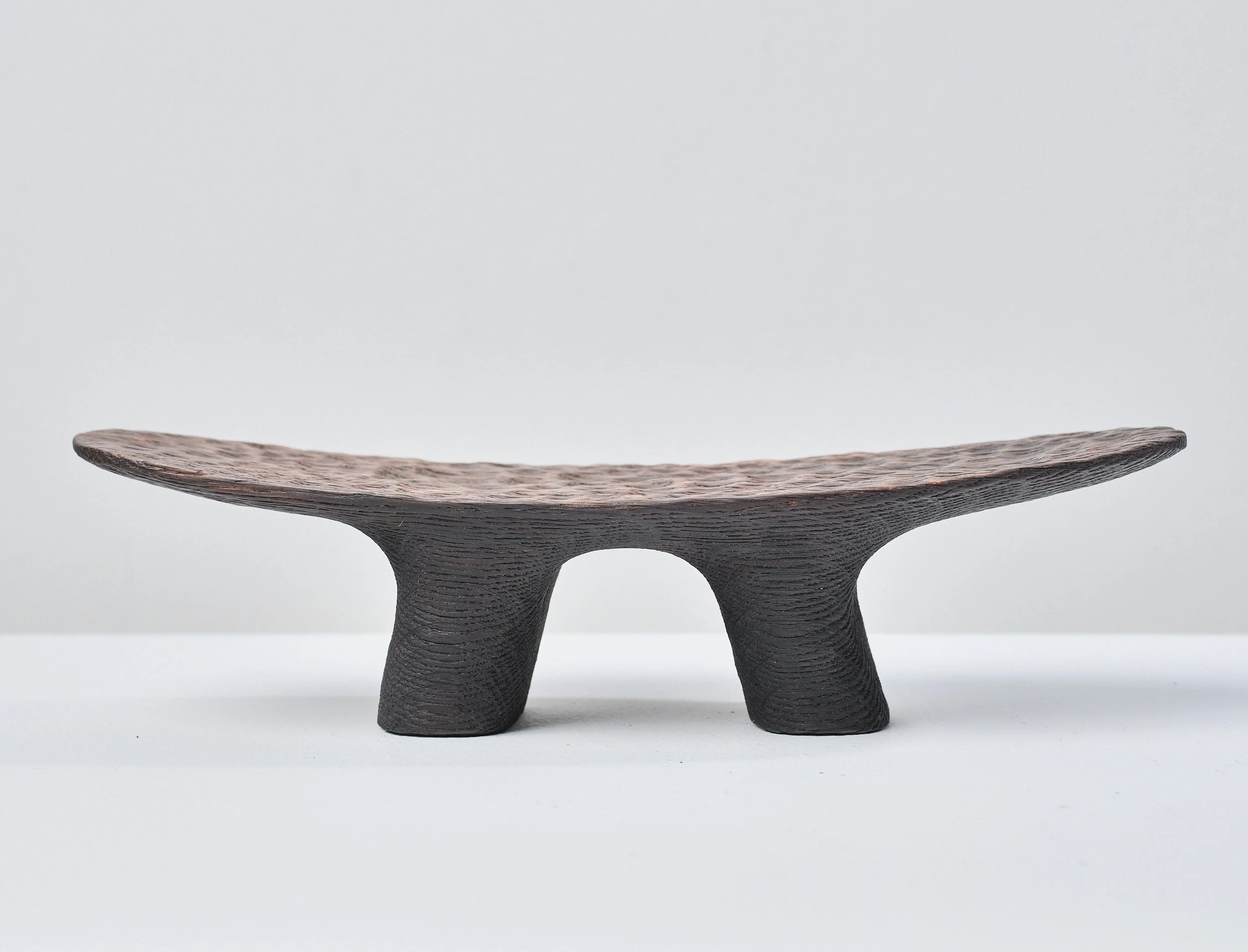 A wooden decorative object with a curved, elongated shape made from blackened oak with polished top, supported by two short, thick legs, placed on a white surface against a plain light background.
