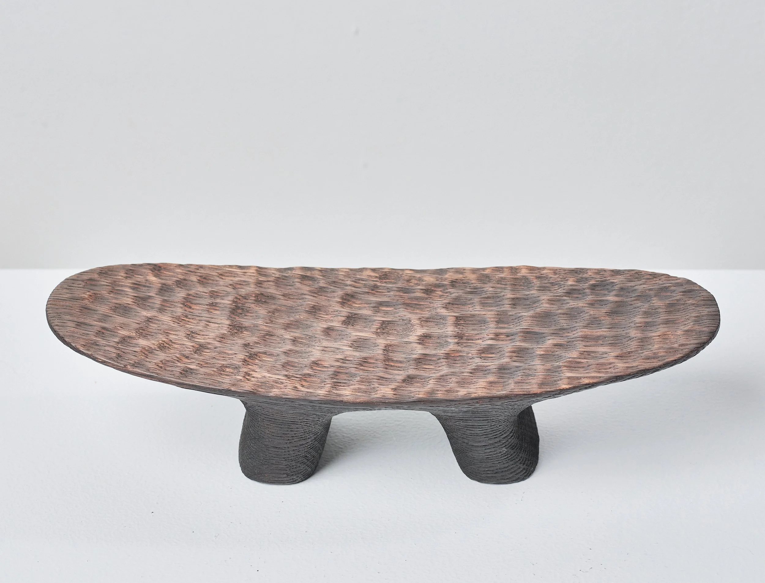 A small, oval-shaped sculpture made from oak from a salvaged beam. The base and legs are charred with heavy grain and the top is dimpled and polished. Sits on a white table against a white wall.