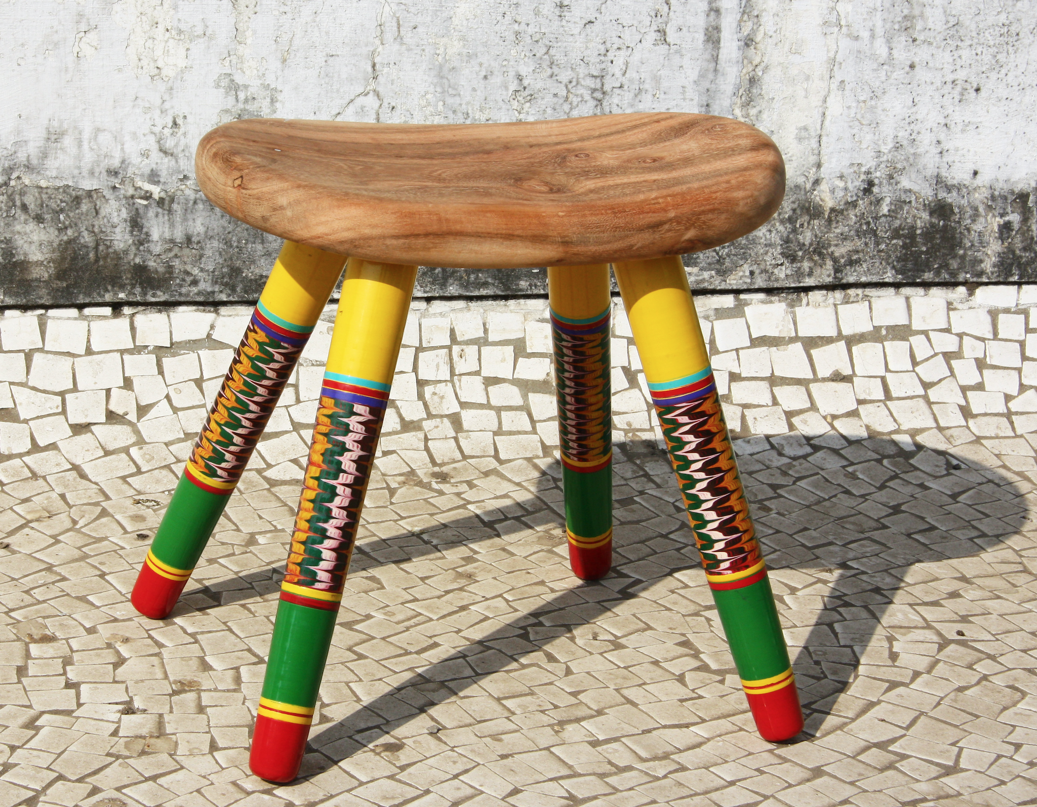 Colourful wooden stool with a natural wood seat and artistically patterned, lacquered legs, placed on a cobblestone pavement against a weathered concrete wall. Part of the Translocated Making research project examining craft interpretations in India