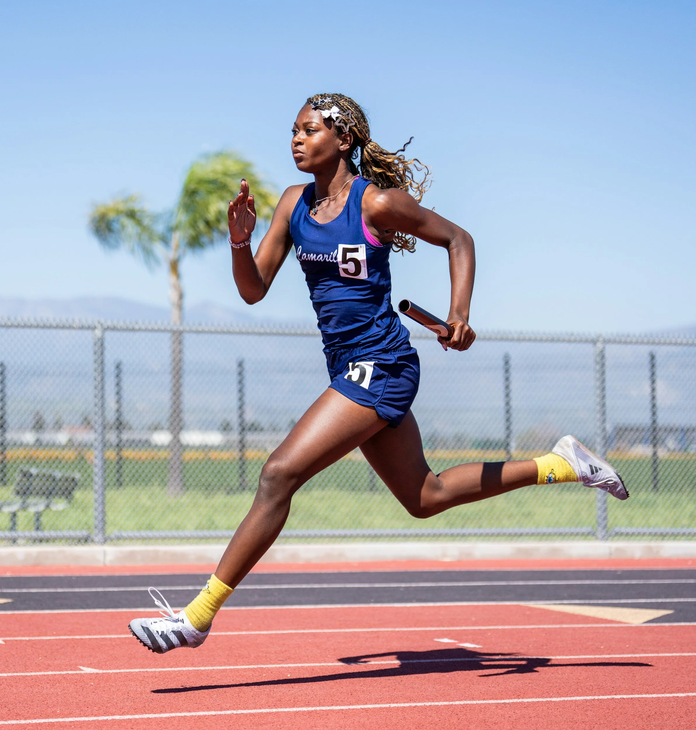 A female athlete is running on a track during a race, wearing a navy blue sports uniform with the number 5, yellow socks, and gray running shoes. She has her hair styled with decorative hair clips.