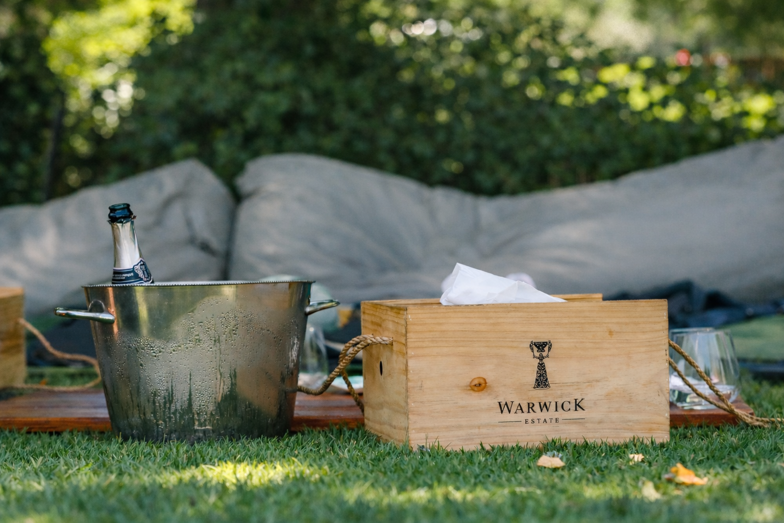 A silver ice bucket with a bottle inside, a wooden box labeled Warwick Estate, a clear stemmed glass, and a wooden surface at an outdoor gathering with greenery in the background.