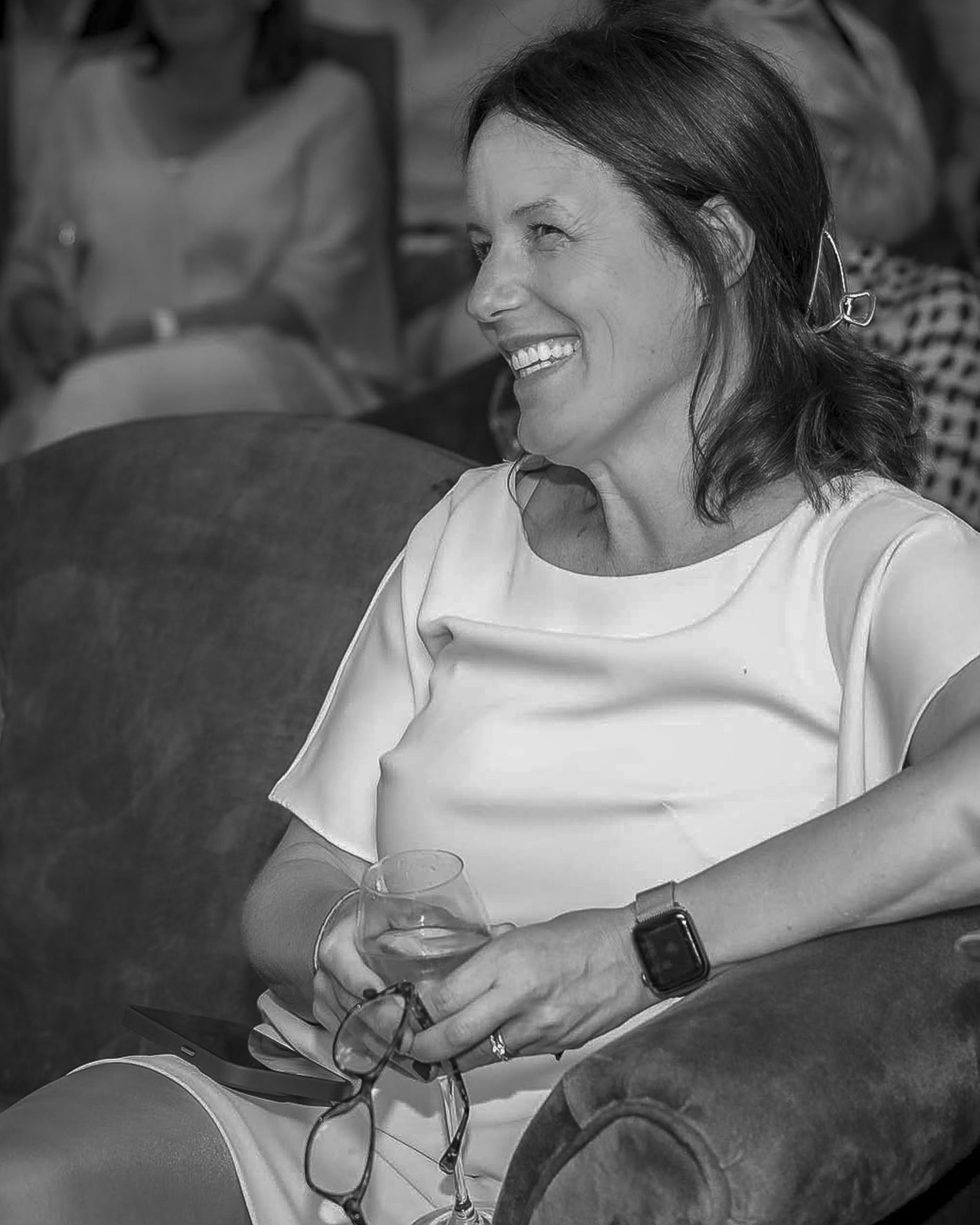 A woman smiling and sitting on a sofa holding a glass of wine, wearing a white top and a smartwatch.