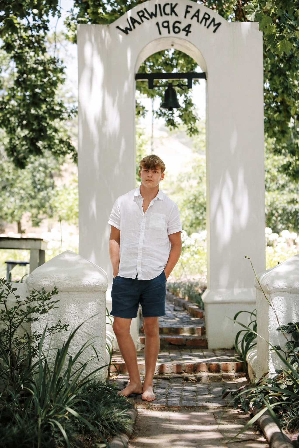 A young man in a white shirt and navy shorts standing barefoot on a brick path at Warwick Farm, under an archway with a bell, surrounded by greenery.