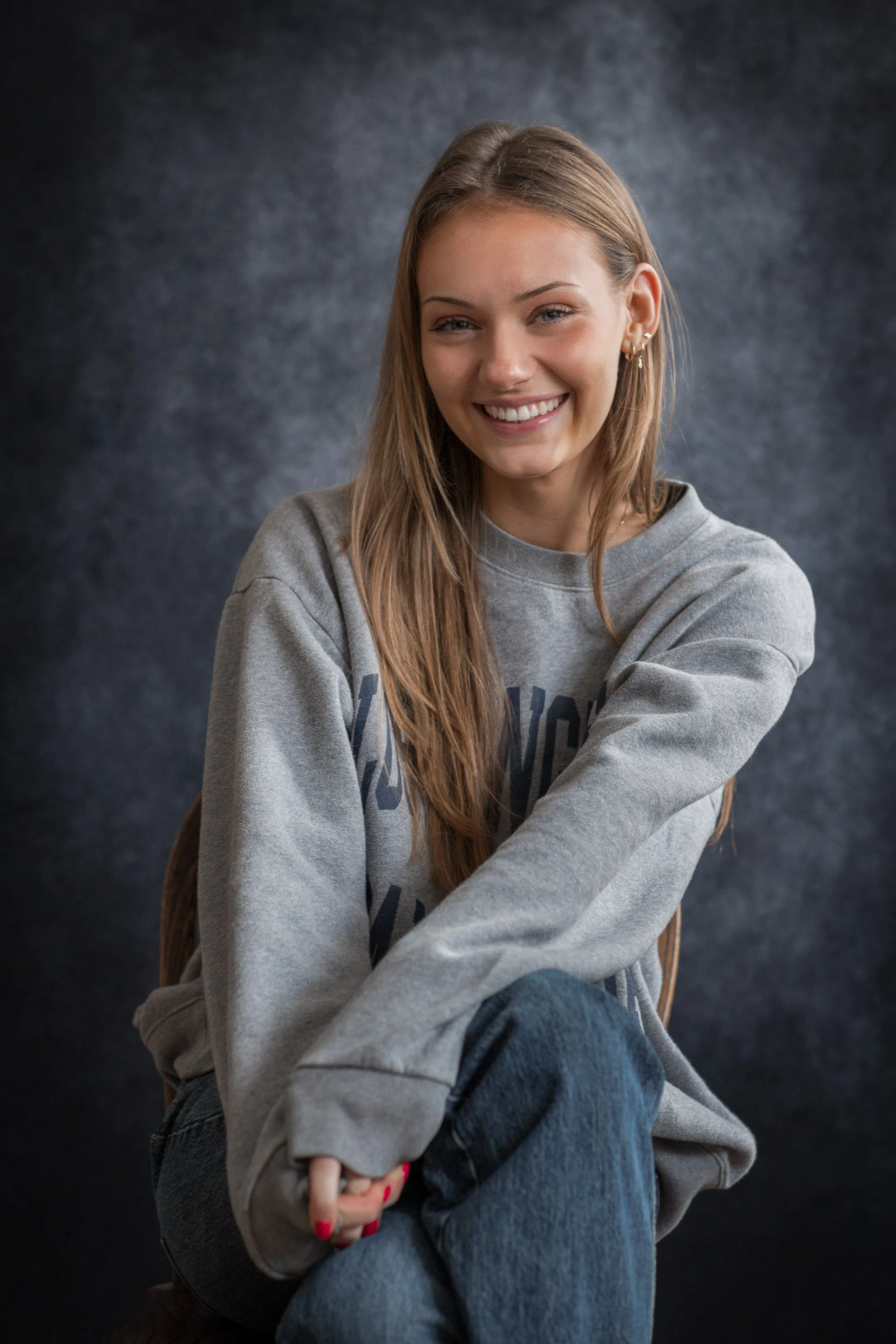 A young woman with long, light brown hair and earrings, smiling while sitting against a dark, textured background, wearing a gray sweatshirt and jeans.