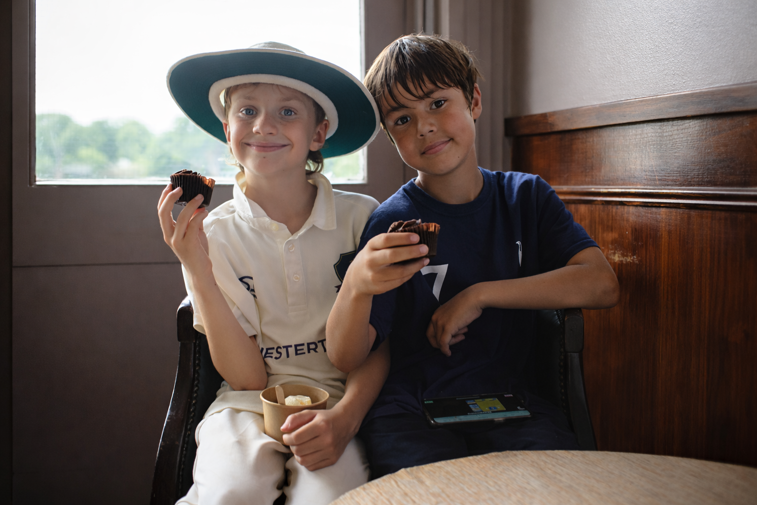 Two boys sitting at a table, enjoying chocolate cupcakes and ice cream, inside a room with a window and wooden paneling.