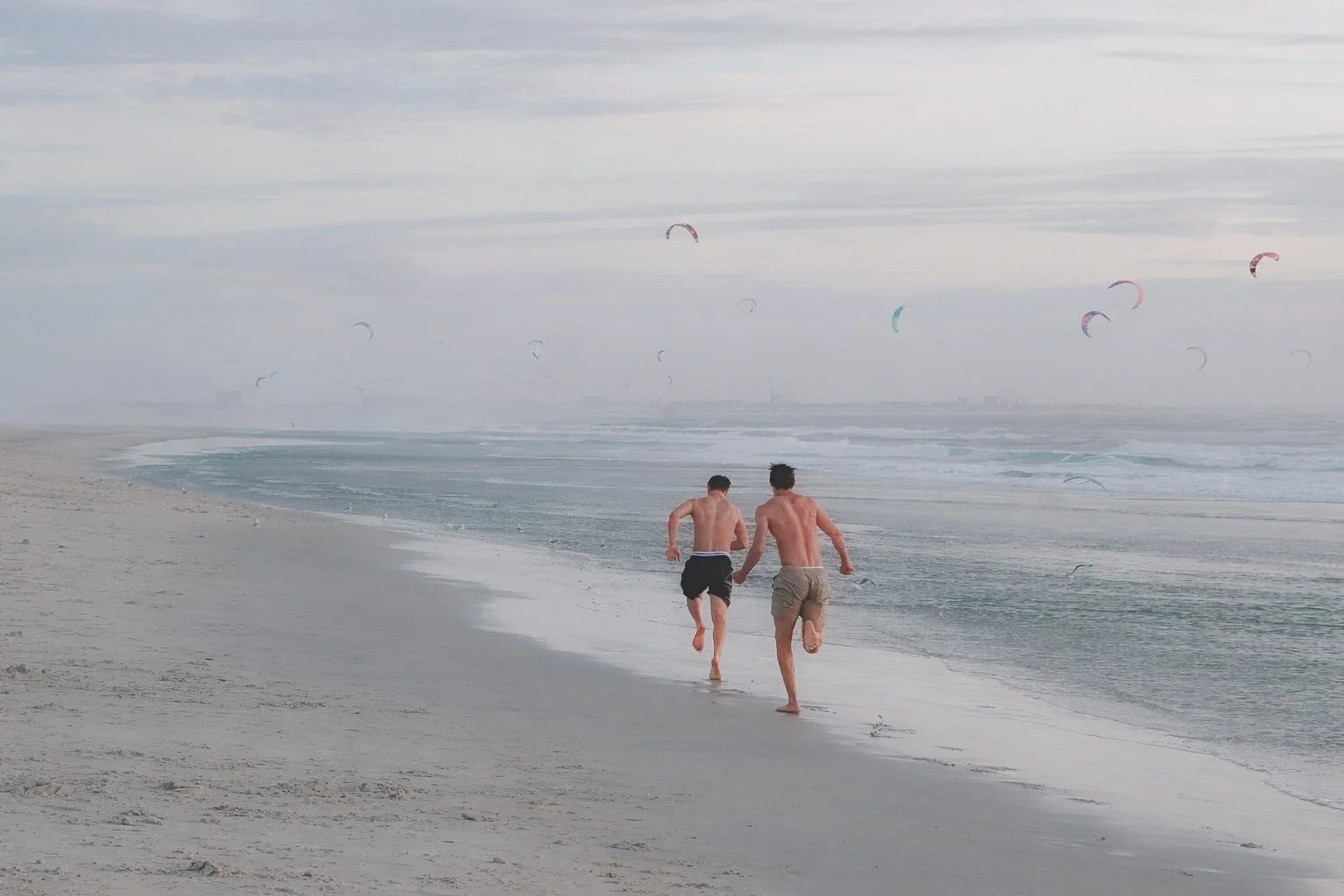 Two young men running on the beach near the shoreline, holding hands, with kite surfers in the sky in the background.