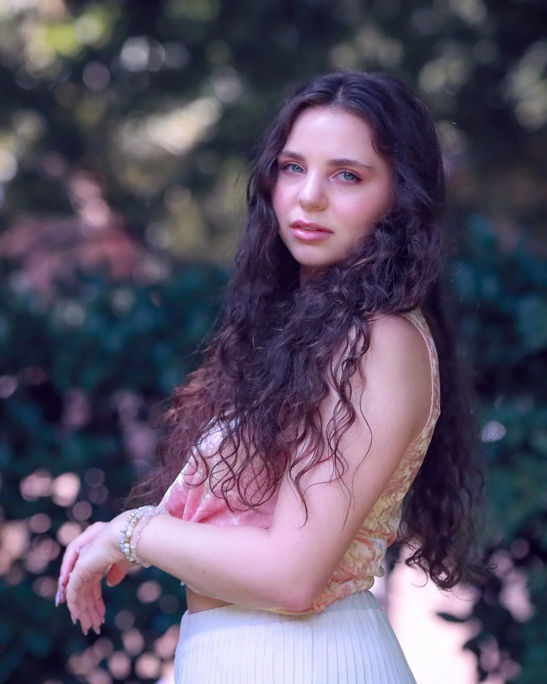 A young woman with long, curly dark hair standing outdoors, looking at the camera with a neutral expression, in a natural setting with blurred foliage in the background.